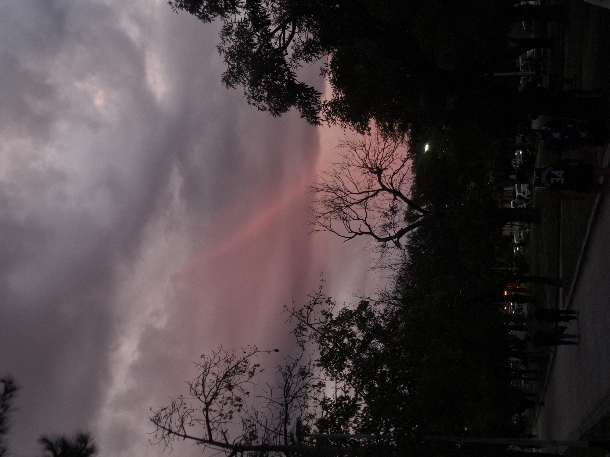 The image depicts an urban park scene at twilight in Villa Domínico, Argentina. The sky dominates the upper frame, characterized by dense, dark purplish-gray clouds, transitioning into a vibrant pink and red hue towards the horizon. A prominent, vertical beam of intense pink light is visible through the clouds in the upper-center. Numerous trees, some bare and silhouetted with intricate branch patterns, others with dark, dense foliage, frame the scene and are largely underexposed against the sky.
In the lower half of the image, a paved pathway extends from the foreground into the mid-ground. Several individuals are present on this path, appearing as dark silhouettes due to the low light. In the foreground, an individual is visible from behind, wearing a dark sleeveless top with the number "34" on the back. Another person walks adjacent to them. Further down the path, additional dark figures are discernible. An illuminated streetlight with a yellowish-white glow is visible in the mid-ground, slightly to the right of the center. Beyond the trees and figures, faint lights from distant vehicles or buildings are visible, confirming an urban setting. The overall lighting suggests late evening or early night.