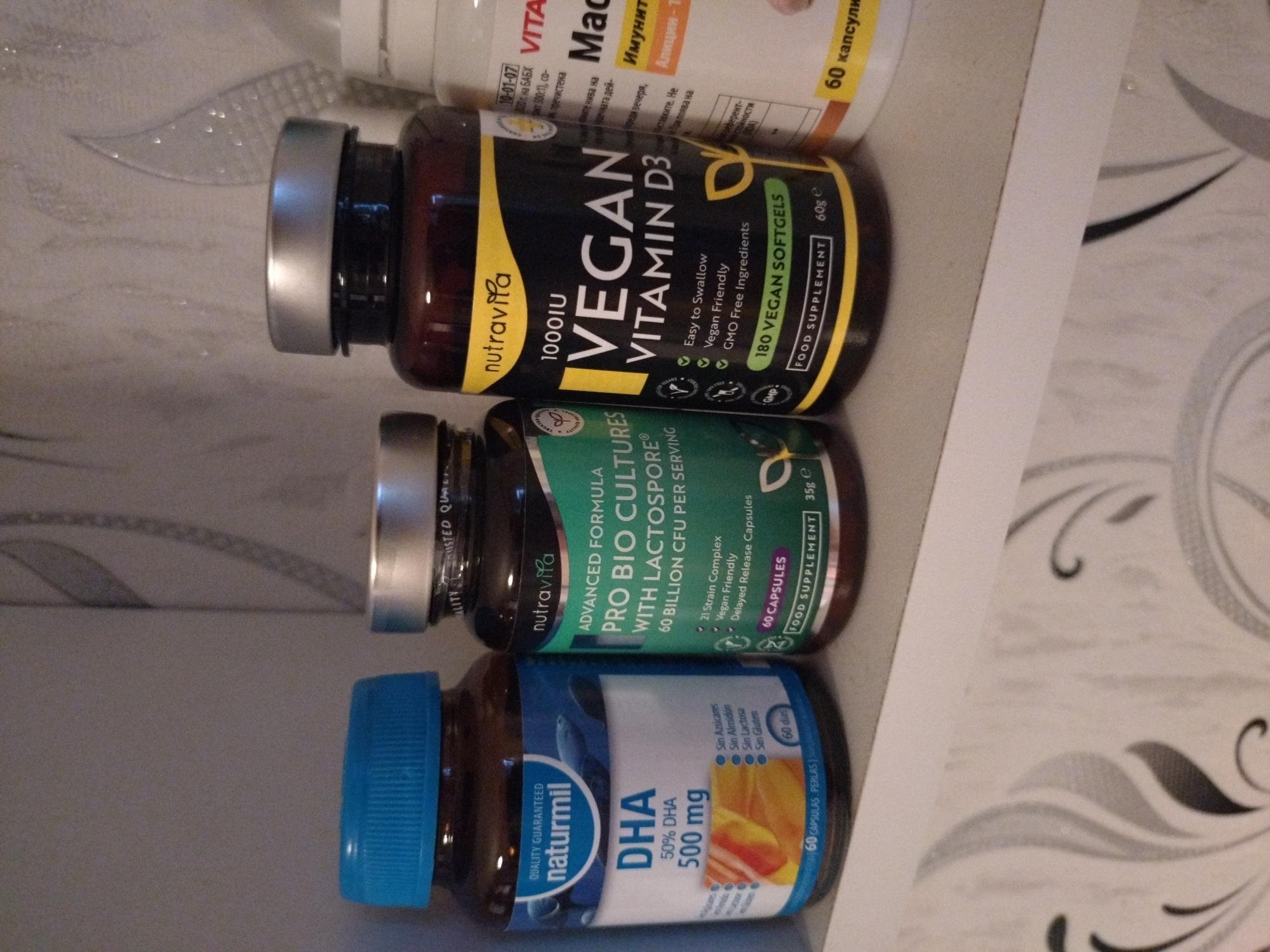 A close-up, eye-level shot shows three bottles of supplements on a shelf. From left to right, the first bottle is dark brown with a blue cap and a white label that reads "naturamil DHA 500 mg". The second bottle is dark brown with a silver cap and a black and yellow label that says "nutravita VEGAN VITAMIN D3 1000 IU". The third bottle is white with a white cap and a label that says "VITA Mac...". The background is a light gray surface with a decorative pattern.