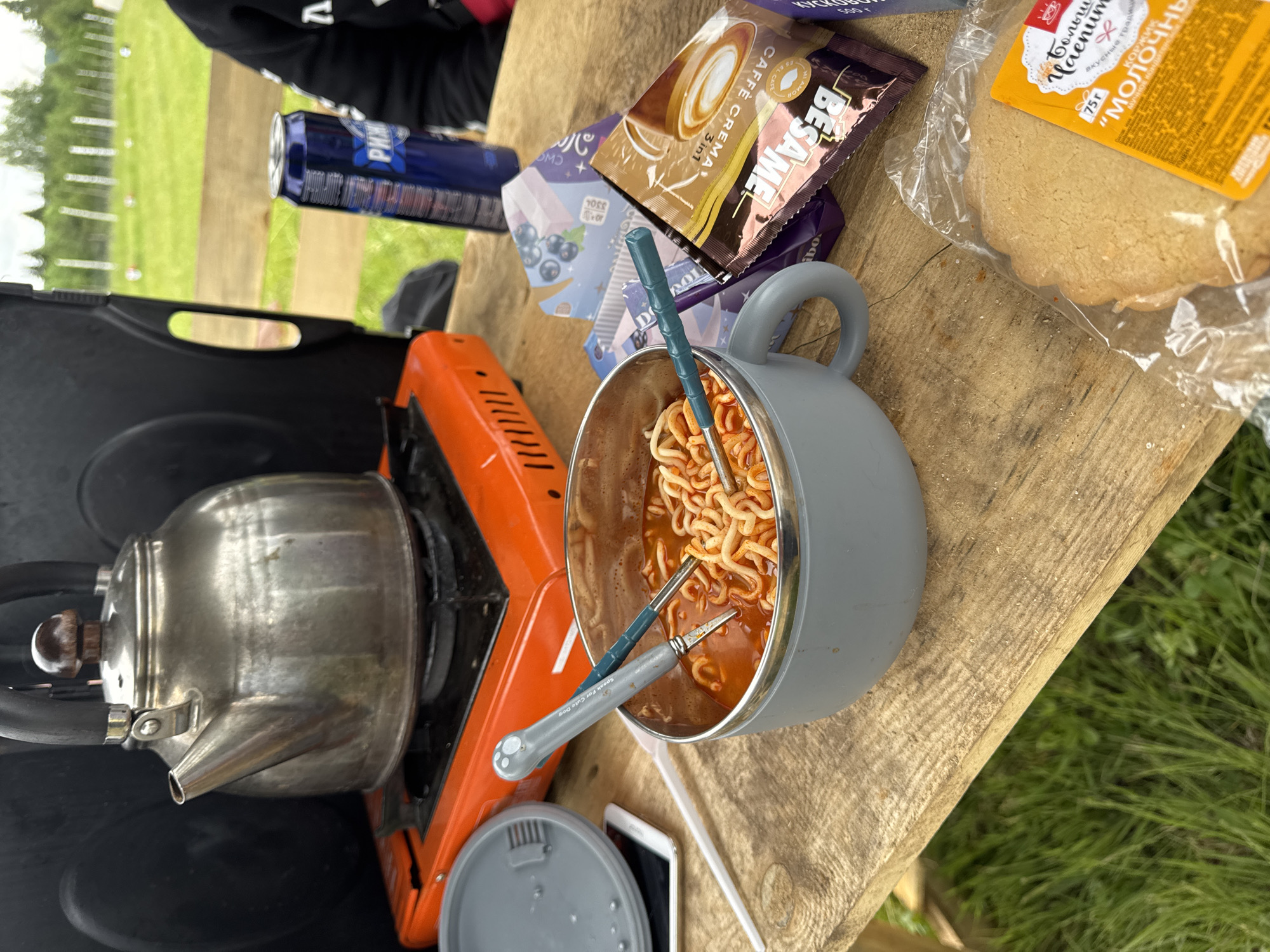 This image captures a casual outdoor meal or snack break, likely during camping or a picnic. The main focus is a grey camping mug or pot filled with instant noodles in a red-orange broth, with two blue and grey collapsible utensils resting inside. One utensil clearly reads "Speak For Cute Dog."
To the left of the mug, a silver metal kettle sits on an orange portable gas stove, suggesting hot water is being boiled. The items are arranged on a rustic wooden surface, possibly a table, which is set against a backdrop of green grass and distant trees, indicating a natural, outdoor setting.
Around the central food items, various provisions are visible. These include sachets of instant coffee labeled "CAFFÈ CREMA 3 in 1" and "BESAME," a packet of "Большой Пряник" (Big Gingerbread/Cookie) which is a "КОРЖИК" (cookie/biscuit) described as "МОЛОЧНЫЙ" (Milky) and weighing "75 г." Other items include a dark blue beverage can (partially visible, showing "PREM") and a purple packet possibly containing sweets, with text like "330 г." A white smartphone and a white straw are also on the table, near a grey lid.
A person, wearing a dark top, is partially visible in the upper left corner. The natural lighting suggests it is daytime. The presence of Russian text on several food packages aligns with the provided location context of Большая Кивара, Russia. The overall scene conveys a relaxed atmosphere of enjoying simple provisions outdoors.