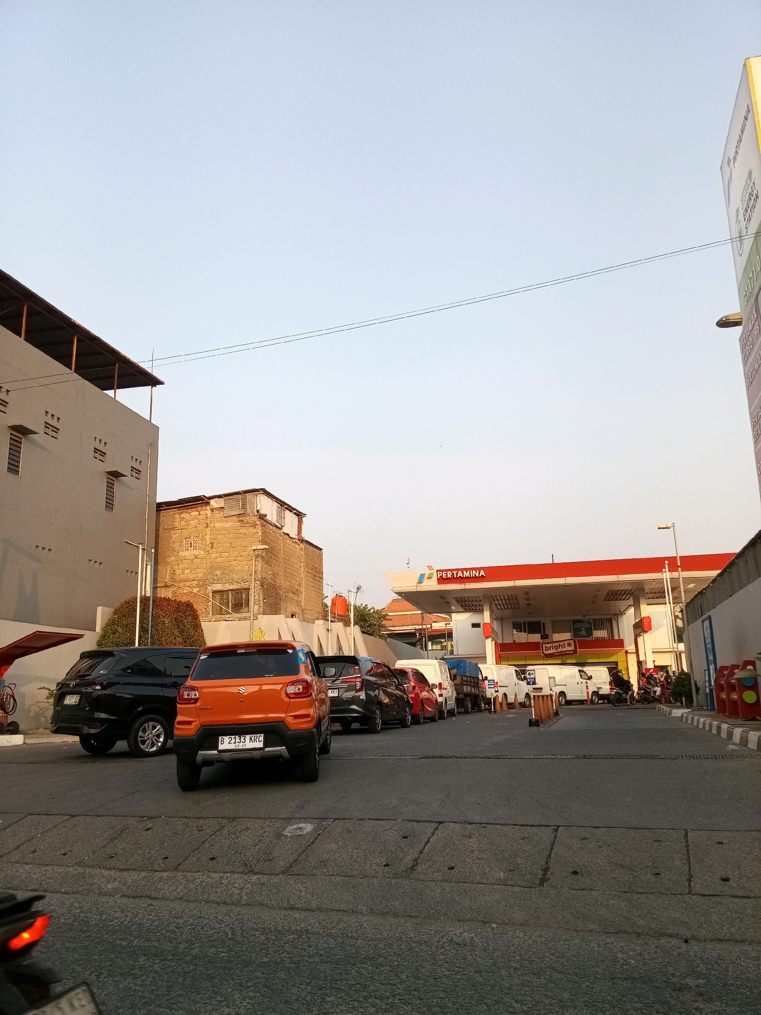 The image shows a street scene in Bekasi, Indonesia, likely a busy area with a Pertamina gas station. Several cars are lined up, suggesting a queue at the gas station. In the foreground, an orange car with license plate "B 2133 KRC" is prominent. Adjacent to it is a black SUV. Further down the street, more vehicles are visible, including white vans and a truck. The gas station itself has a red and white canopy with the "PERTAMINA" logo displayed. Buildings are visible on either side of the street, one appearing to be a multi-story structure with windows and vents, and another made of brick. The sky is clear and pale blue, indicating daytime. The overall environment appears to be an urban setting, typical of a roadside commercial area.
