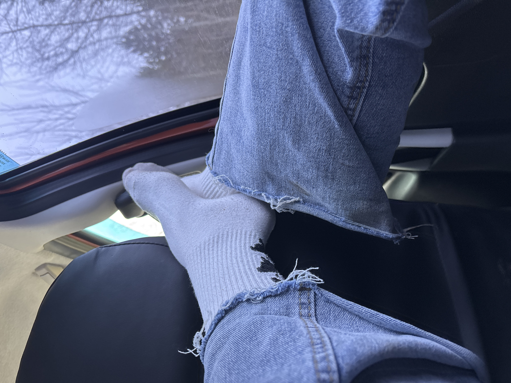 A pair of legs clad in distressed light blue denim jeans and white socks are elevated inside of a vehicle. The feet rest on the upper frame of what appears to be a hatchback or trunk opening. The interior features a dark, possibly leather, seat and a light-colored headliner. Through the vehicle's window, a snow-covered landscape with bare trees is visible. The image provides no indication of being in Dubrova, Russia.