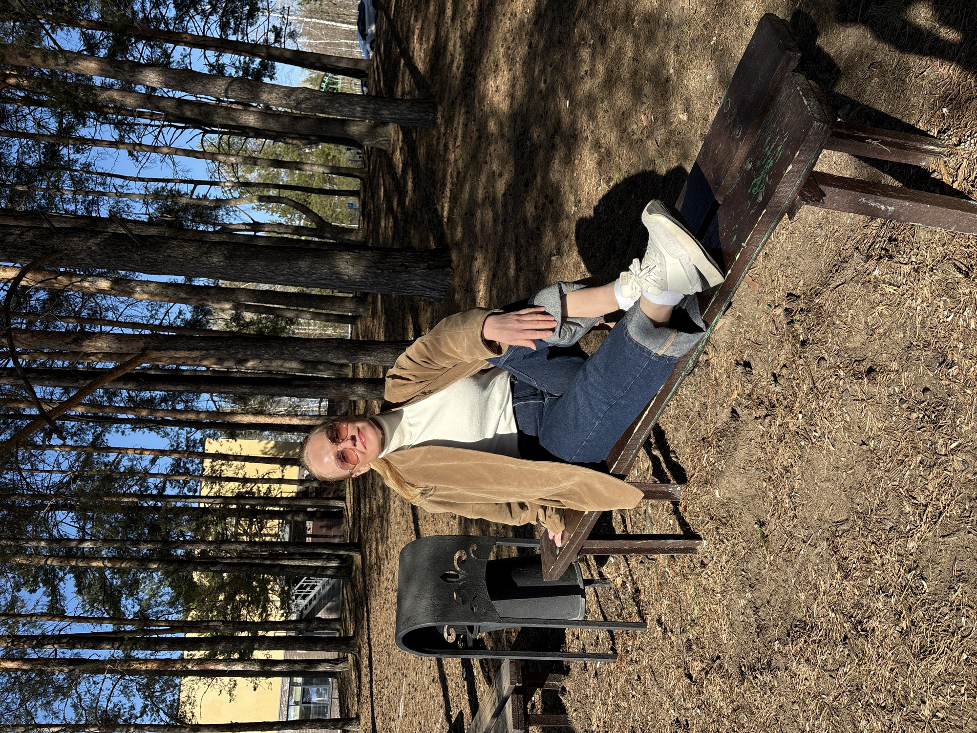 The image captures a full-body view of a person relaxing on a wooden bench in a sunny pine forest, presumably in Chaikovsky, Russia. The shot is rotated ninety degrees counter-clockwise.
A young person, possibly a woman, is casually reclining on a dark brown wooden bench, facing slightly towards the right side of the frame. She is dressed in a white turtleneck sweater, a light brown/tan jacket, dark blue jeans with rolled-up cuffs, and white sneakers with white socks. Her blonde hair is pulled back, and she is wearing light-tinted sunglasses, looking towards the viewer with a relaxed, possibly smiling, expression. Her left hand rests on her leg, while her right arm is bent, with her hand near her hip.
In front of the bench, a black metal trash bin or urn with decorative scrollwork cutouts is visible. The ground is covered with dry pine needles, fallen leaves, and patches of earth, characteristic of a forest floor. Tall, slender pine trees with rough, dark bark dominate the scene, casting long shadows on the sun-drenched ground, indicating a bright, clear day, likely midday or early afternoon. Through the trees, a vibrant blue sky is visible, and in the distant background, parts of light-colored buildings, possibly with yellow facades and windows, can be seen, along with a partially obscured white vehicle. Some illegible light-colored text or graffiti is faintly visible on the backrest of the wooden bench. The overall atmosphere is peaceful and relaxed.