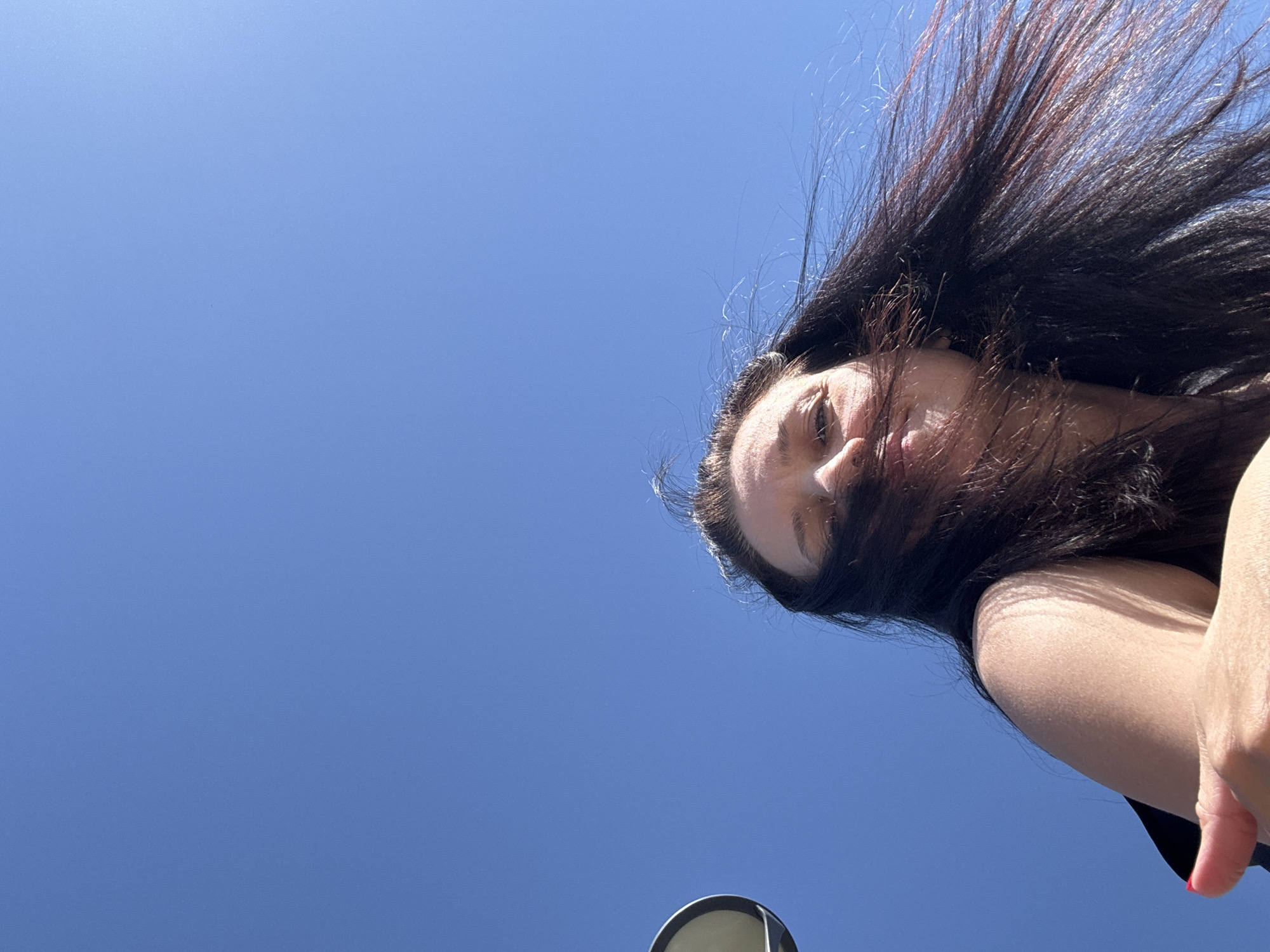 A low-angle shot captures a person with long, dark hair against a clear blue sky. The hair is windblown, obscuring part of the face. The person's nose and part of their eye are visible. The person is wearing a dark shirt. A portion of an arm and hand is visible in the lower right corner. In the bottom center of the frame, a blurred, round object is present. The location is indiscernible.