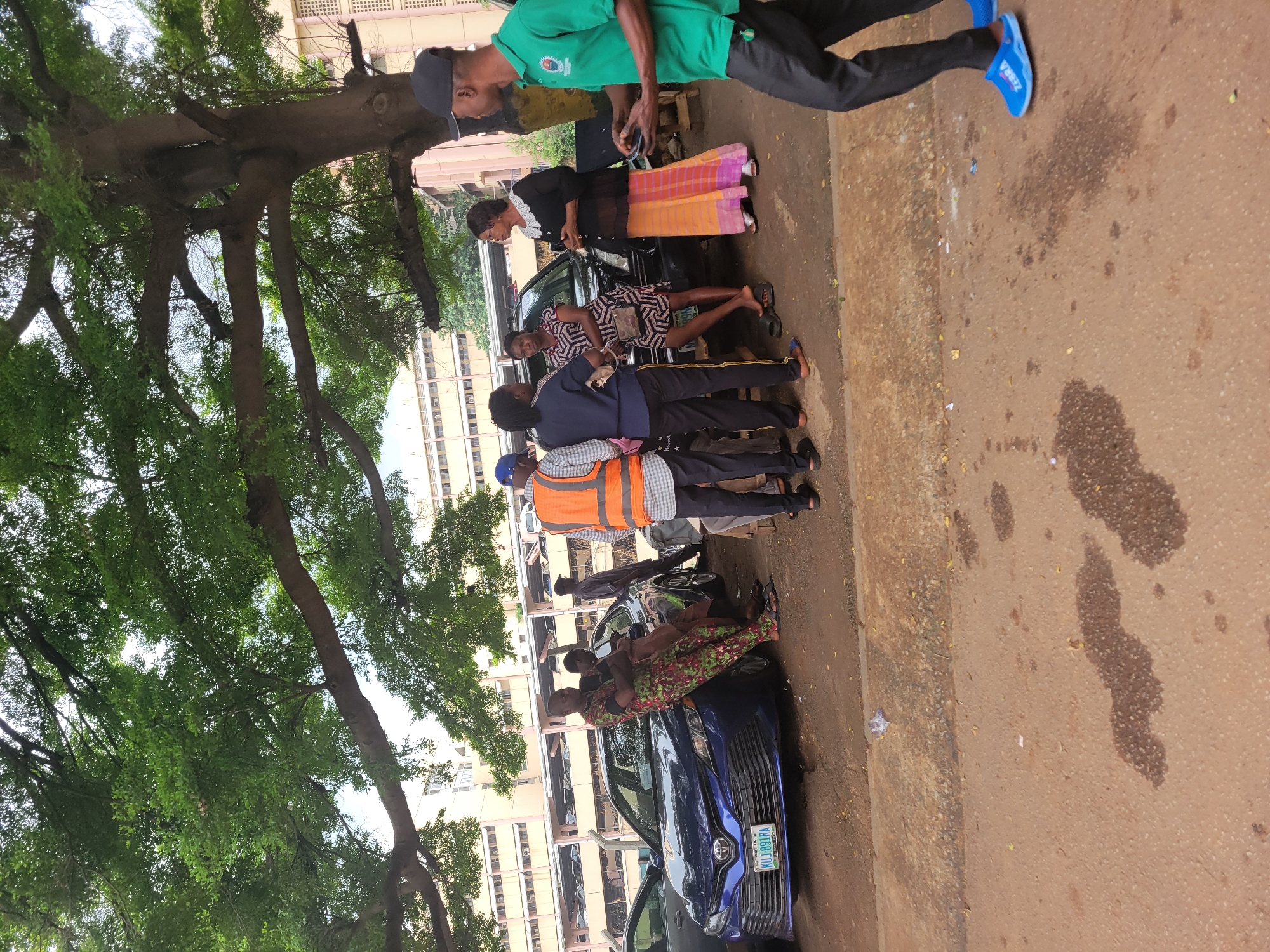 The image depicts an outdoor scene in Abuja, Nigeria, under a large tree with dense green foliage. In the foreground, a reddish-brown paved surface with dark wet patches is visible. A group of individuals are gathered near two parked cars. A blue sedan, a Toyota Camry with a license plate reading "KUJ-891RA", is prominently positioned. To its right is a darker SUV.
Several people are interacting. One man, wearing a vibrant green shirt and dark pants, is walking away from the viewer towards the right of the frame, holding a smartphone. He is wearing blue clogs. To his left, a woman in a black top and a long, brightly colored orange and pink skirt is speaking with a group of people standing near the SUV. A woman in a patterned dress is also part of this group. Another man in an orange high-visibility vest and a plaid shirt is standing nearby. Further to the left, two individuals are leaning against the blue sedan, one in a colorful patterned outfit and the other in a brown garment. The background shows a multi-story building with a grid-like facade, likely part of an urban infrastructure. The overall impression is a casual gathering or interaction in a public space.