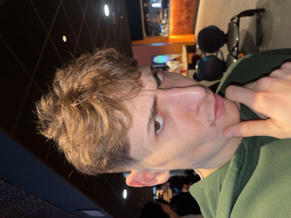 A young man with short, wavy, light brown hair poses indoors, his head angled. His eye is hazel-colored, and he has fair skin with a slight stubble. He wears a green hooded sweatshirt. His right hand is raised, with his index finger touching his chin. Behind him is a dark, textured wall and, indistinctly, a blurred bar-like setting with lighting fixtures and other people. Sunglasses are visible hanging on a shelf in the background.