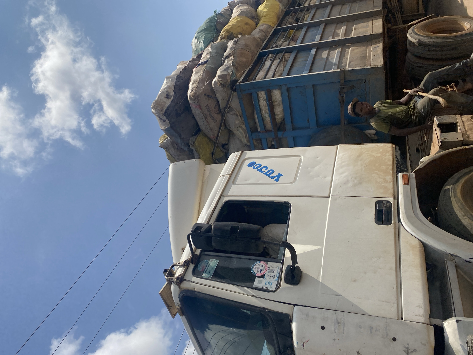 A full shot captures a white truck loaded with large sacks, parked on a dirt road under a bright blue sky with scattered white clouds. A young man, wearing a green t-shirt and dark pants, sits on the side of the truck, holding a shovel-like tool. The truck's cabin is visible on the left, with a side mirror and stickers on the window. The rear of the truck is a blue, open-sided trailer packed with various sacks of different colors and sizes. The ground is dusty, and power lines are visible against the sky. The scene appears to be in Ado, Nigeria.