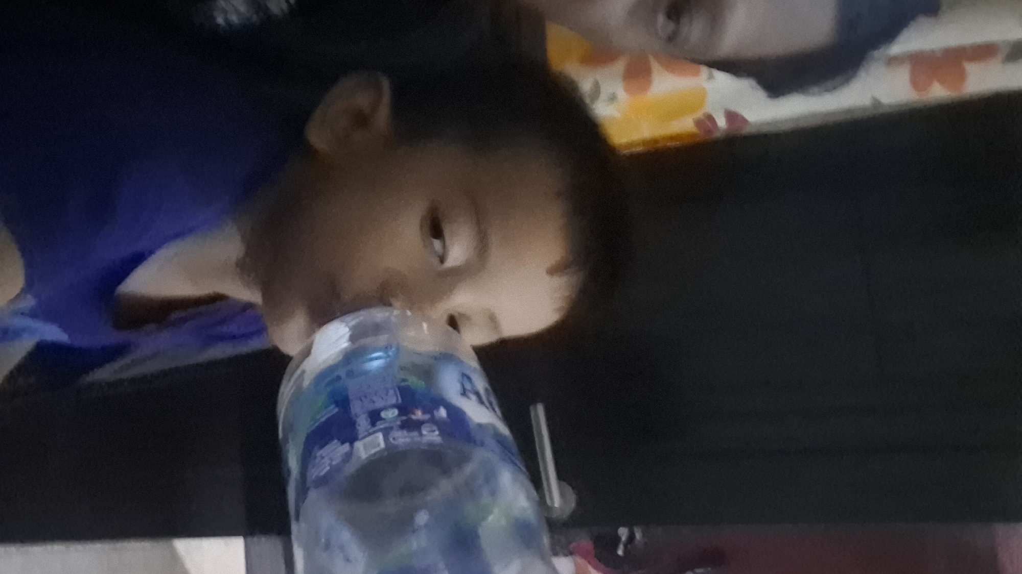 A young child is drinking from a clear plastic bottle of water. The child is facing right and has dark hair and eyes. The child is wearing a blue shirt. In the background, there is a dark wall with a silver door handle. To the left of the child, a curtain with a floral pattern is visible. The scene appears to be indoors.