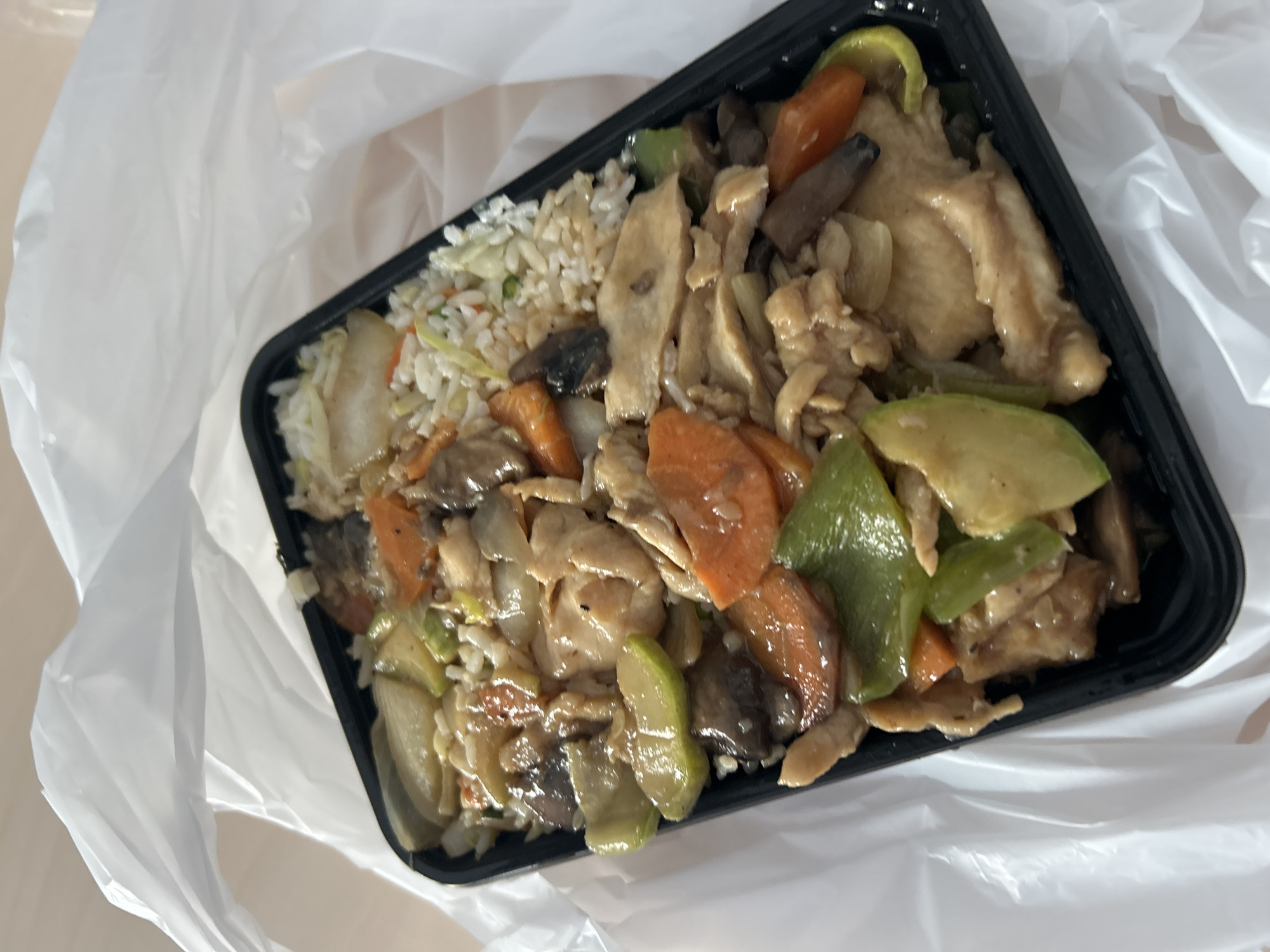 A take-out container of Asian food sits in a white plastic bag. The container is black and rectangular, filled with rice, chicken, carrots, green peppers, zucchini, and mushrooms. The location is unknown, but is not necessarily in Sofia, Bulgaria.