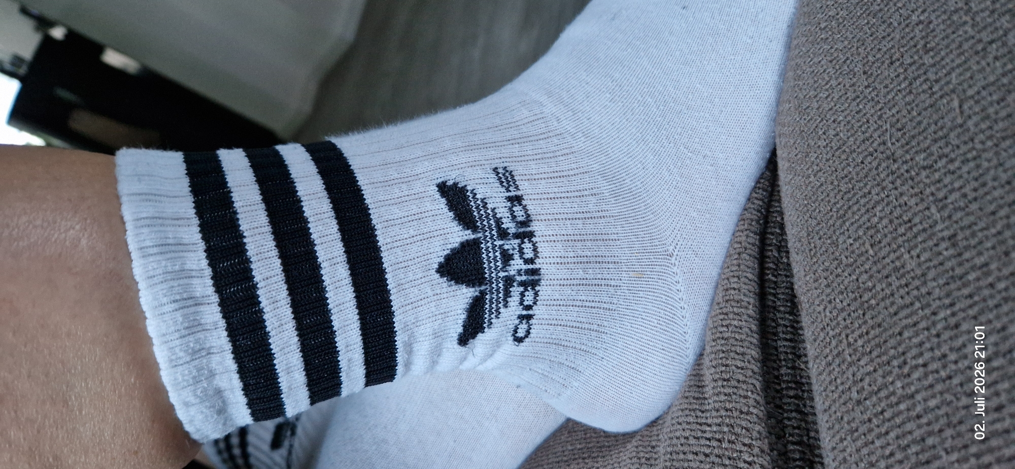 A close-up, low-angle, full shot shows a person's right foot wearing a white ankle sock with black Adidas branding and three black stripes near the cuff. The sock is positioned on a brown, textured surface, possibly a couch or blanket. The background is blurred, showing a dark, indistinct object on the right and a lighter surface on the left. A timestamp in the lower-left corner reads "02. Juli 2026 21:01". The image does not provide enough information to determine the specific location within Hummeltal, Germany.