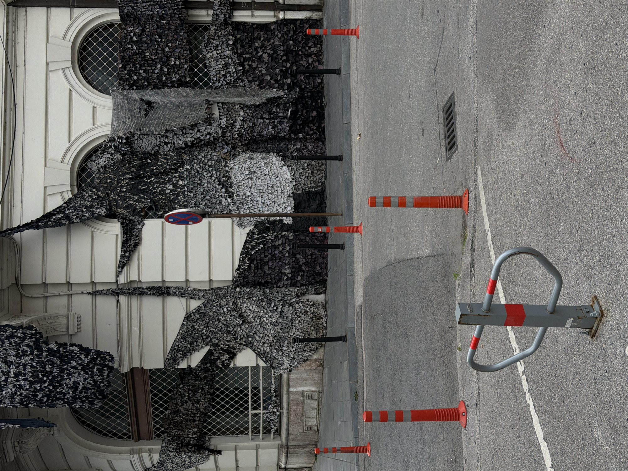 The image depicts an outdoor urban scene, likely in Sarajevo, Bosnia and Herzegovina, given the artistic installation and architectural style. A building with classical architectural elements, featuring arched windows with diamond-shaped grilles, is partially covered by a large, dark, textured art installation. This installation appears to be made of numerous small, irregular pieces of fabric or material, creating a layered, almost organic form that drapes over the building's facade.
In the foreground, a paved road surface is visible, marked with a white line. Several orange traffic cones with reflective white bands are strategically placed on the road and sidewalk. A metal parking barrier, painted gray with red reflective bands, stands in the foreground. A storm drain grate is also visible on the road. The overall impression is one of an urban environment that has been temporarily transformed by a public art installation.