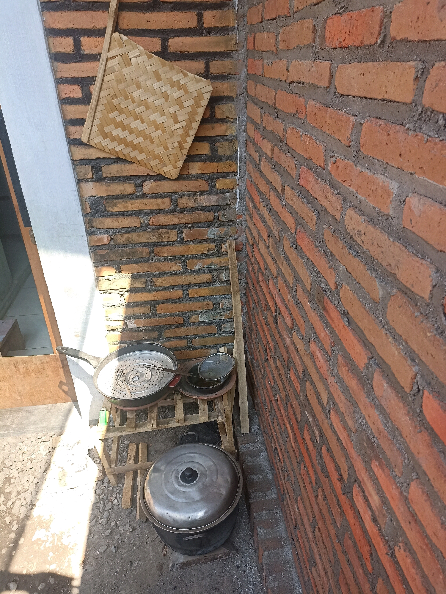 The image depicts an outdoor corner of a building with a brick wall and a doorway. On the brick wall, a woven bamboo fan is hanging. In the foreground, an assortment of cooking utensils is arranged on a wooden rack. These include a large, shallow pan with a perforated insert and a fork, a smaller sieve, and a large pot with a lid. The ground is covered in gravel and dirt, with sunlight casting shadows. The location is identified as Mojopanggung, Indonesia.