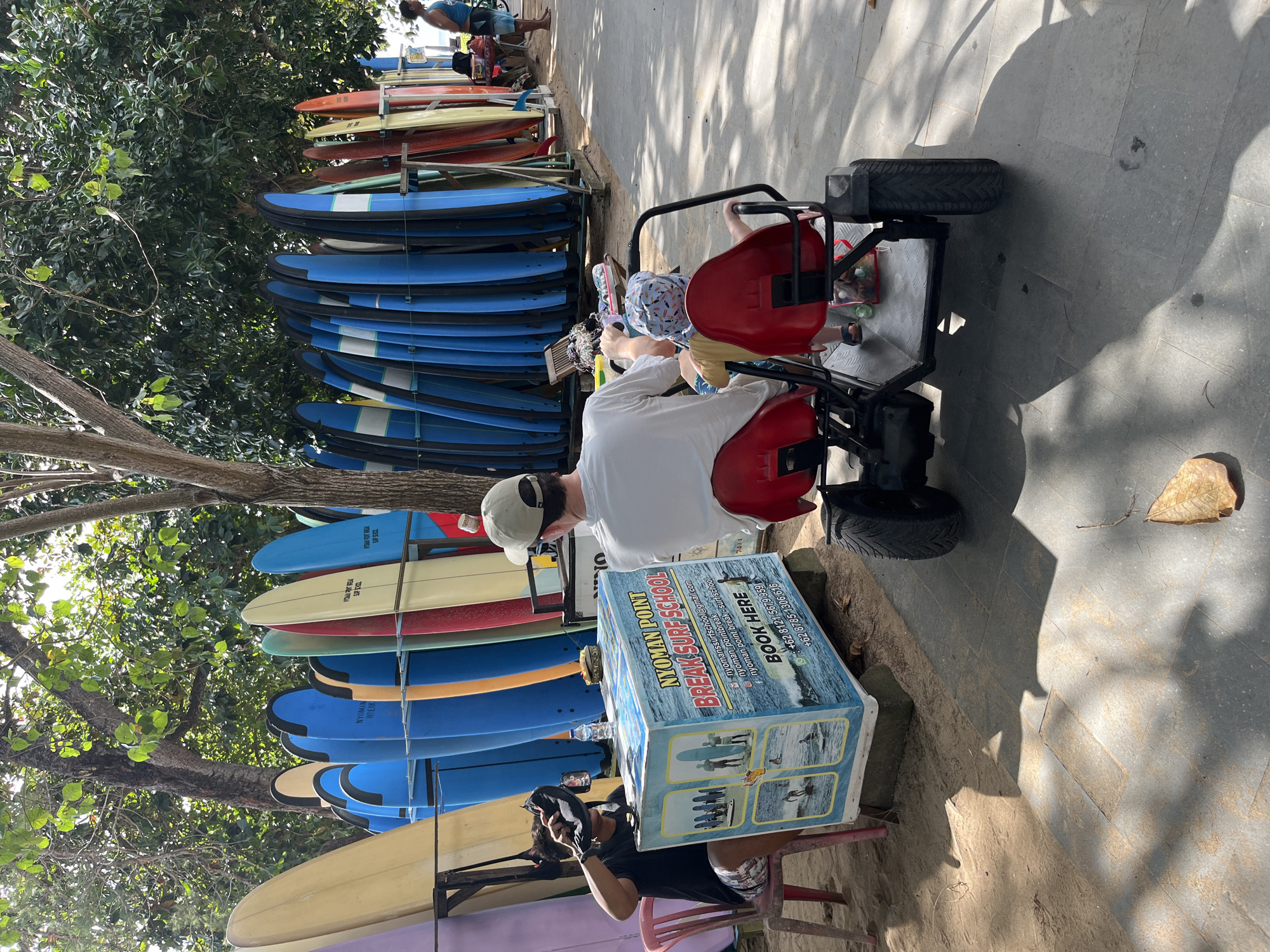 In Kuta Selatan, Indonesia, a row of surfboards stands upright on a beachside promenade. A man sits on a red chair, holding a black hat. In front of him, a blue and white surf school sign displays images of surfing lessons and contact information. A person wearing a white shirt and a beige cap drives a red pedal-powered go-kart with two children seated behind them, moving along the paved path. Another man stands in the background near more surfboards. The environment is sunny with dappled light filtering through trees, casting shadows on the ground.
