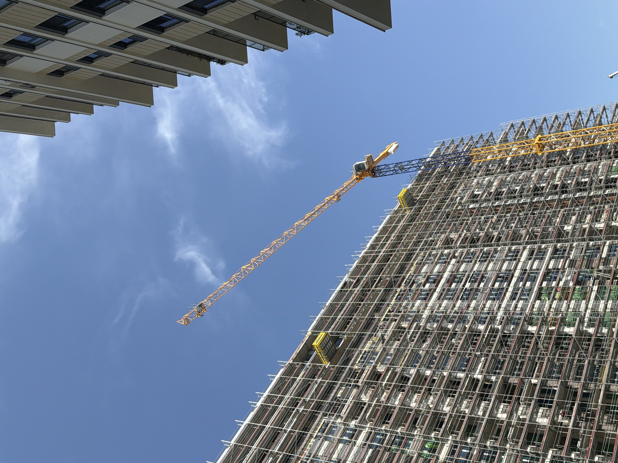 The photograph shows a construction site in Belgrade, Serbia, with a tall building under construction. A construction crane is visible in the center of the frame. The building is covered in scaffolding. Another building is partially visible in the upper left corner. The sky is blue with some wispy clouds.