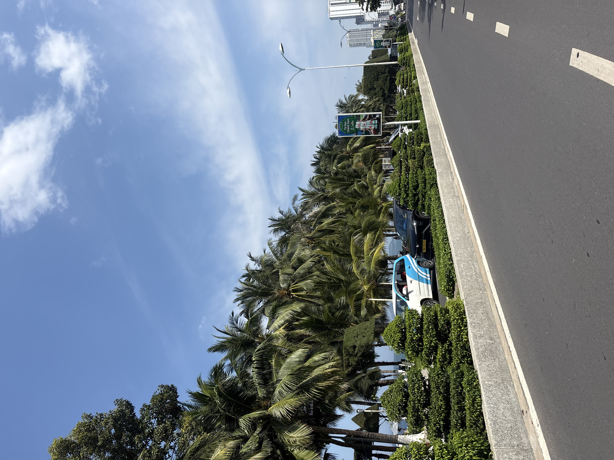 This image depicts a street scene in Nha Trang, Vietnam, characterized by a clear blue sky with scattered white clouds. A paved road runs diagonally from the lower right to the middle left of the frame, bordered by a grey concrete curb and a well-maintained green median strip featuring manicured bushes and palm trees. Several vehicles are visible: a white van with blue accents and a black car are parked or driving on the right side of the road. In the background, tall buildings line the horizon, suggesting an urban environment. A billboard featuring what appears to be a beverage advertisement stands prominently in the median. The presence of lush palm trees and the general atmosphere suggest a coastal or tropical urban setting, consistent with Nha Trang. There are no discernible people in the immediate foreground or engaging in any specific actions.