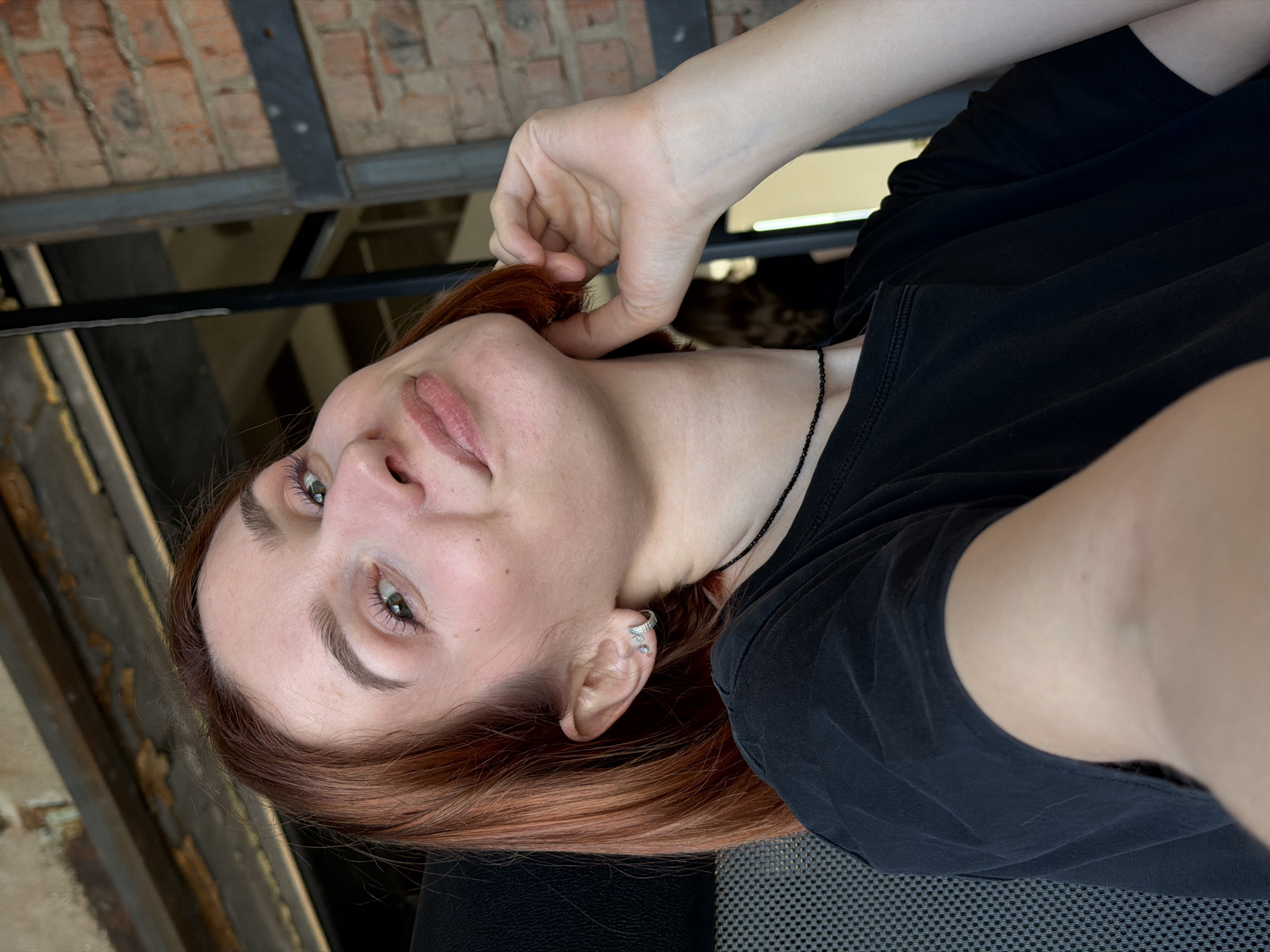 A woman with reddish-brown hair and fair skin is captured in a rotated close-up selfie. She is wearing a black t-shirt and a thin black necklace. She has a silver earring. Her hand is near her face, with fingers lightly touching her hair. The background includes a brick wall and black metal beams, suggesting an urban industrial or commercial location, and could be a cafe or restaurant with interior design elements. The photo was taken in Chaikovsky, Russia.