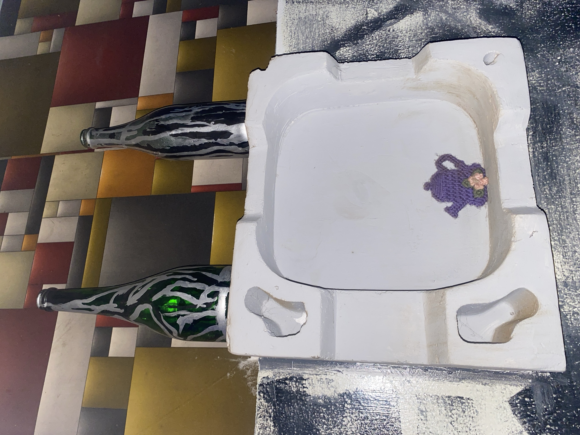 Two decorative bottles, one green with white abstract markings and the other dark with white abstract markings, stand on a white shelf. The shelf is a molded, white, rectangular tray with raised edges and two indentations on the left side, resembling stylized feet. In the center of the tray, a small, crocheted purple teapot with a pink flower sits. The background features a wall with a geometric pattern of squares and rectangles in varying shades of red, gold, and gray. The image appears to be an indoor scene. There are no people visible. The location of Kabong, Nigeria is not discernable from the visual information provided.