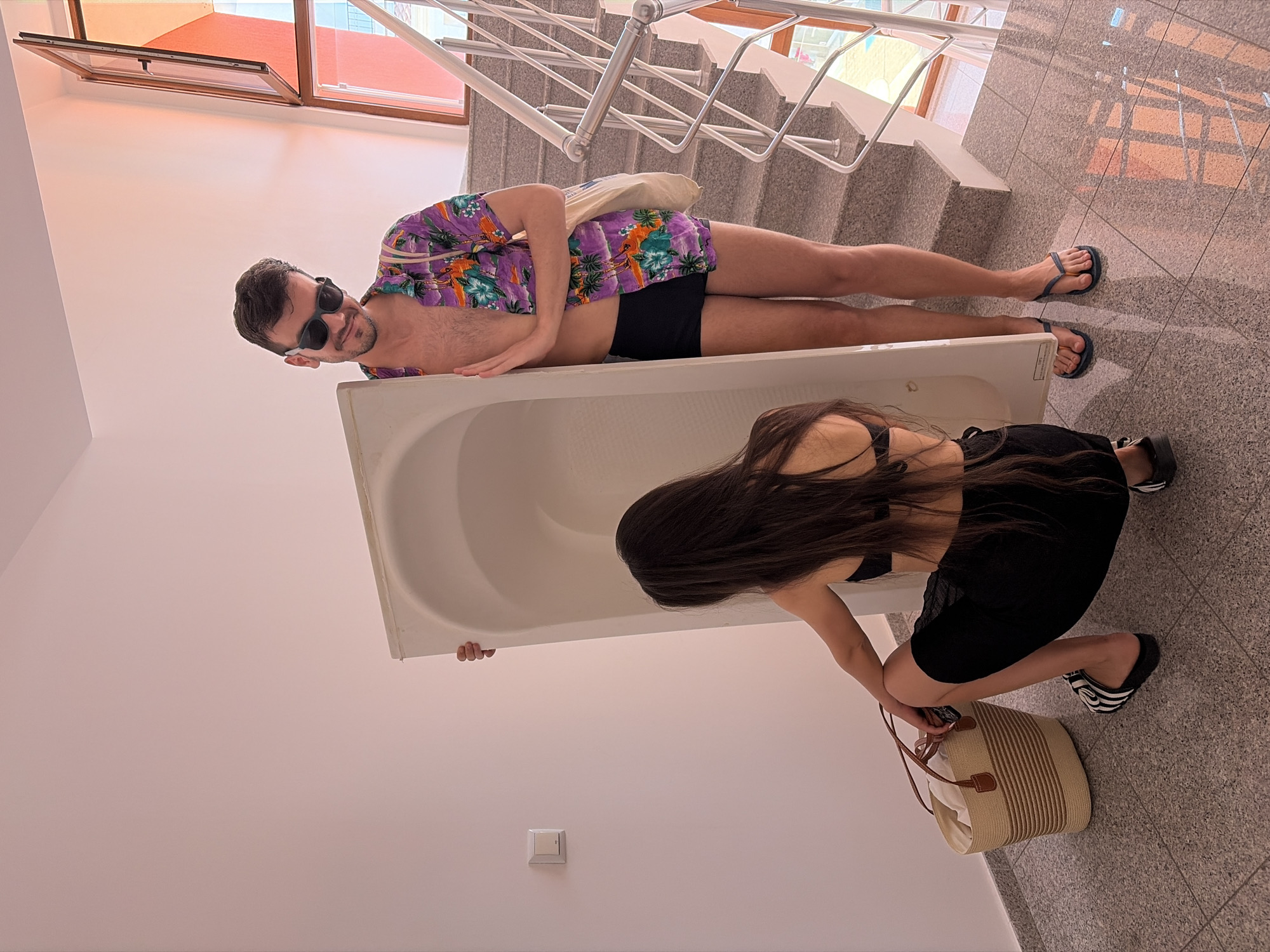 The image shows a man and a woman moving a bathtub. The man, wearing sunglasses, a Hawaiian shirt, and black swim trunks, is lying on top of the bathtub. He has a bag slung over his shoulder. The woman, wearing a black top and skirt, is kneeling beside the bathtub, holding onto it. She has long dark hair and is wearing black sandals. A straw bag with white contents is placed on the floor near her. The bathtub is white and appears to be made of plastic or enamel.
They are on a tiled staircase, which is an unusual place to be moving a bathtub. The lighting suggests it is daytime, possibly indoors or in a shaded outdoor area, as there are reflections on the floor from what appears to be a window. The overall scene is somewhat surreal and humorous due to the context of moving a bathtub on a staircase with the man lounging on top. There are no visible signs or text in the image.