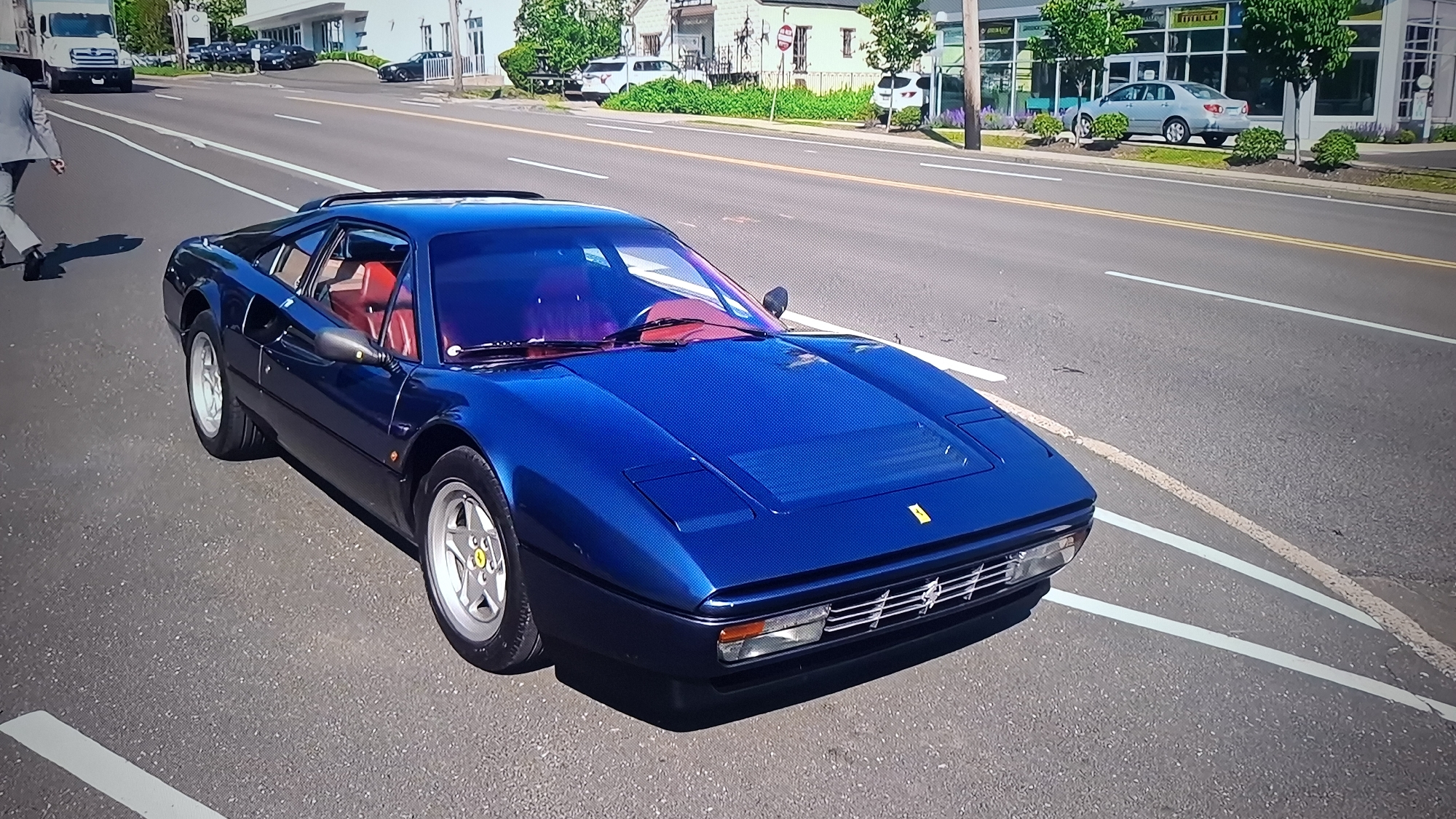 A blue Ferrari 308 GTS sports car is parked at an angle in the roadway with red interior visible. A man in a light-colored suit is walking on the road next to the car. Buildings line the other side of the roadway with other cars visible. Trees and landscaping are present.
