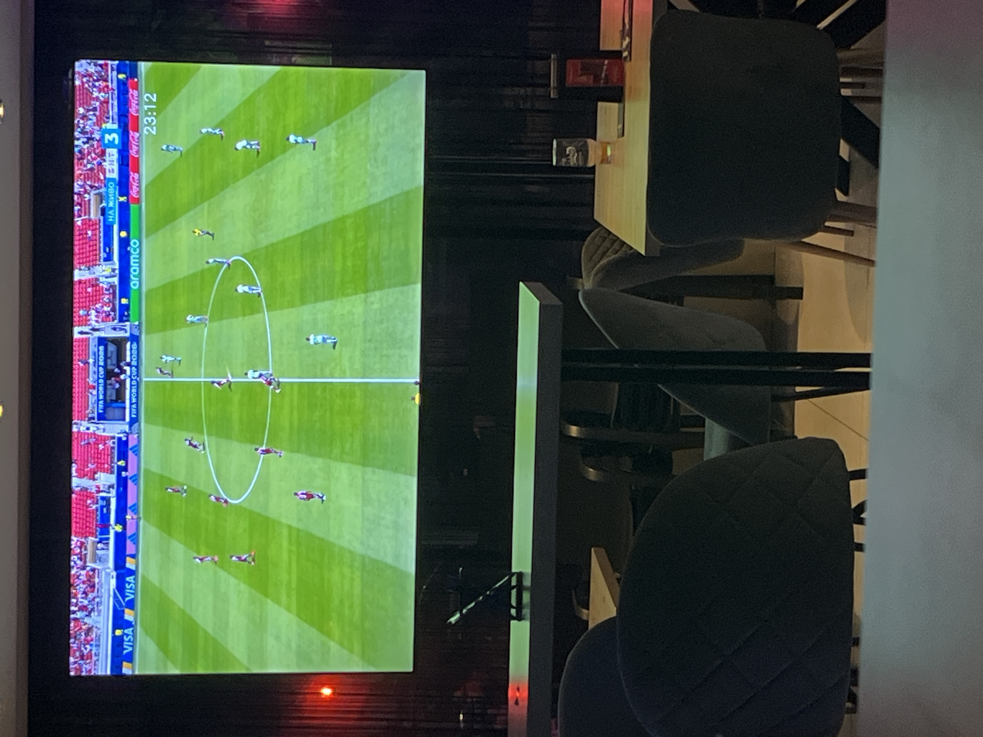 An indoor scene depicts a large screen displaying a soccer match. The screen shows a top-down view of a soccer field with players in red and white uniforms, a referee, and advertising banners along the sidelines, including VISA, FIFA World Cup 2026, Aramco, and Coca-Cola. A digital clock on the screen shows "23:12". In the foreground, a table with chairs is visible, suggesting a viewing area. A partially filled glass of beer sits on the table. The lighting is dim, with red accent lighting on the left side of the screen.