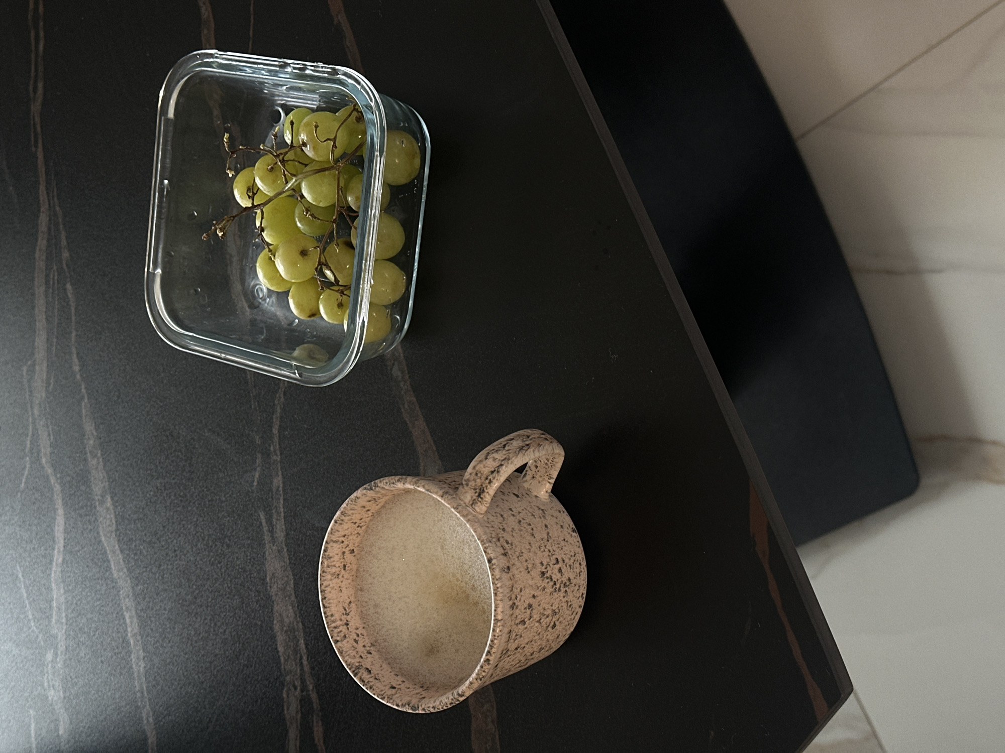 A high-angle, medium shot depicts a glass container filled with green grapes and a speckled, light-brown mug on a dark, marbled tabletop. The table's surface has lighter gray streaks, creating a textured appearance. A dark, possibly black, piece of furniture is partially visible in the upper right, along with a marble-patterned floor or countertop. No people are visible, and no actions or interactions can be inferred from the image. The location is unknown, but since the user asked it is in Sofia, Bulgaria.