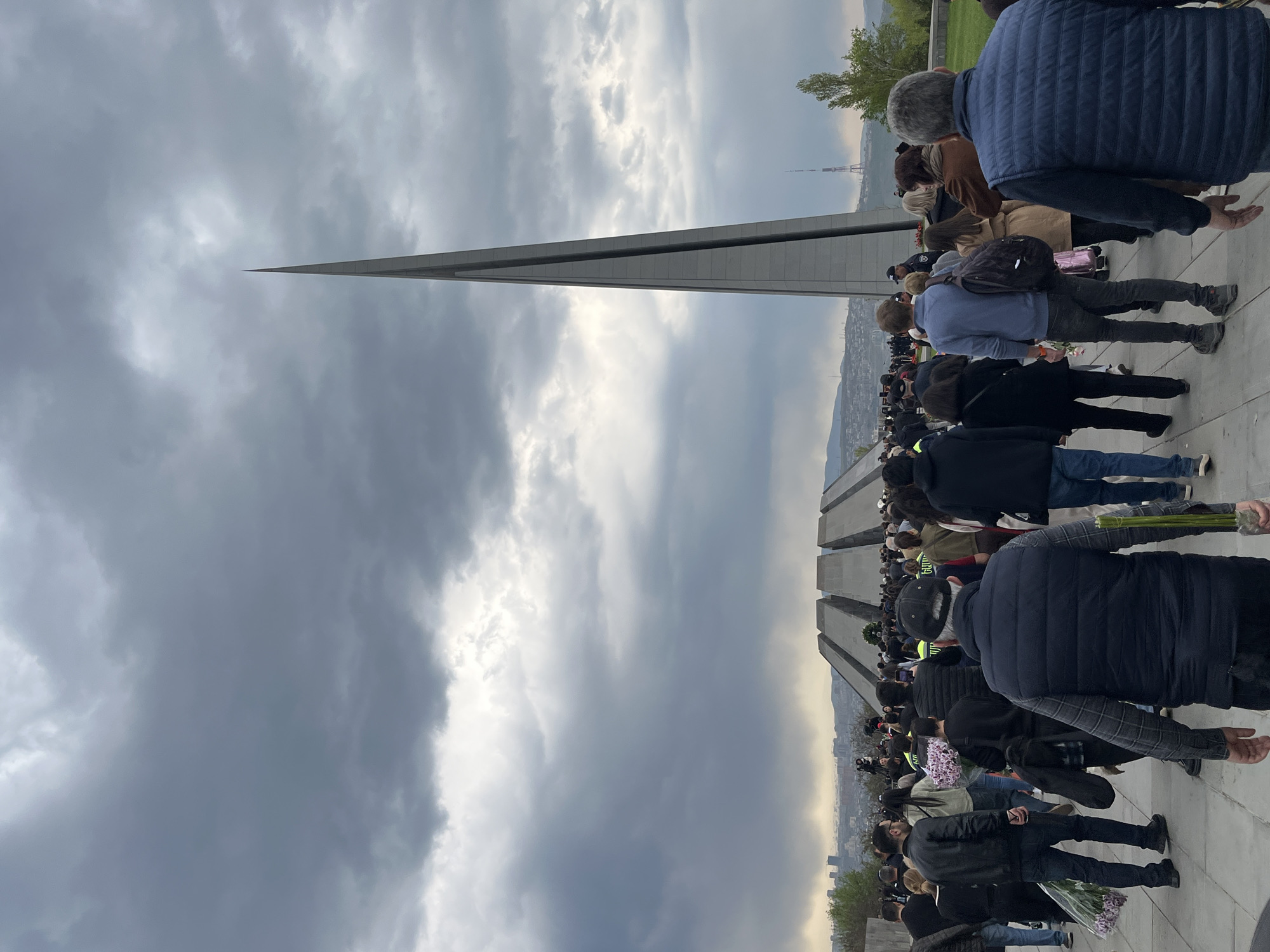 The image, originally rotated 90 degrees counter-clockwise, depicts a solemn gathering at an outdoor memorial complex under a cloudy sky. A large crowd of people is visible, mostly from behind, forming a line or moving towards a central point. Many individuals are carrying bouquets of flowers, suggesting a commemorative event or an act of tribute.
In the center-left of the correctly oriented image, a tall, slender, pointed monument, resembling an obelisk, dominates the skyline. To the right, in the mid-ground and background, several large, grey, trapezoidal stone structures, characteristic of memorial stelae, are arranged. This architecture, combined with the context, identifies the location as the Tsitsernakaberd Armenian Genocide Memorial Complex in Yerevan, Armenia.
The sky is heavily overcast with grey clouds, though some brighter areas indicate daylight, possibly late afternoon. The overall atmosphere appears somber and respectful. In the distant background, a cityscape stretches towards distant hills, and a communication tower is faintly visible. People are dressed in various casual attires, including jackets and vests. One person in the crowd wears a yellow high-visibility vest with faint text that appears to be "ՈՍՏԻԿԱՆՈՒԹՅՈՒՆ" (Armenian for "POLICE"). The scene captures a moment of collective remembrance or mourning.