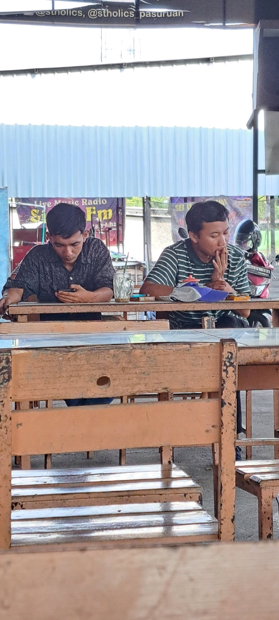 Two young men are seated at a wooden table outdoors, with a corrugated metal roof overhead and a light blue corrugated metal wall in the background. The man on the left, wearing a dark patterned shirt, is looking down at his phone. The man on the right, wearing a green and white striped shirt, is smoking a cigarette. A glass with a straw, a pack of cigarettes, and a blue baseball cap are on the table between them. A red and white motorcycle is visible behind the man on the right. A sign reading "Live Music Radio FM" is in the background. The scene appears to be in a casual outdoor eating or seating area.