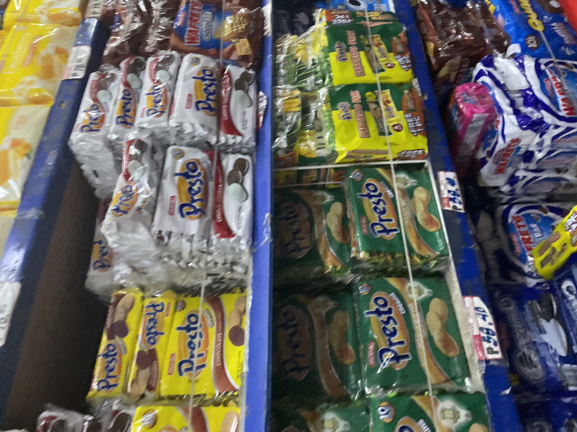 The image displays a detailed view of a retail store shelf in Davao City, Philippines, primarily featuring packaged biscuits and snack items. The scene is illuminated by overhead lighting, casting minimal shadows and providing clear visibility of the products.
The topmost visible shelf, bordered by a blue metal trim, contains various packaged goods, predominantly in yellow and white packaging, though specific brands are largely out of focus. A white price tag displaying "P 76.00" is affixed to the blue trim, indicating the Philippine Peso currency.
The main focus is on the middle shelf, which is well-stocked with "Presto" brand biscuits. On the left, three yellow packages of "Presto" chocolate-coated biscuits are stacked vertically, each displaying a dark biscuit partially covered in chocolate. In the center, multiple white and silver packages of "Presto" biscuits are stacked; these feature circular biscuits, some showing white cream filling and text "choco vanilla" discernible on one pack. To the right of these "Presto" stacks, brown and reddish packages of "Wafrets Choco Bar" are visible, appearing as individually wrapped wafer bars.
A blue shelf ledge separates the middle and lower sections. A prominent white price tag on this ledge clearly displays "P 59.40."
The lower shelf, visible through a wire grid, showcases more packaged goods. On the left, several green packages labeled "Presto Creamies" are stacked, depicting light-colored oval biscuits. Adjacent to these, yellow packages marked "Presto Favorito Pack" are stacked, with the text "FREE 1 PC" also visible on their labels. Further to the right, similar yellow "Favorito Pack" items are seen, alongside blue-packaged snacks, possibly "Oreo" brand or similar, stacked with a small yellow toy car resting on top.
Below this, a partially visible lowest shelf contains additional stacks of white and blue packaged "Wafrets" and "Wafer" products, with a small pink package resting on top of a stack of the white "Wafrets." The overall environment suggests an organized retail display within a grocery or convenience store setting. No people are visible in the frame.