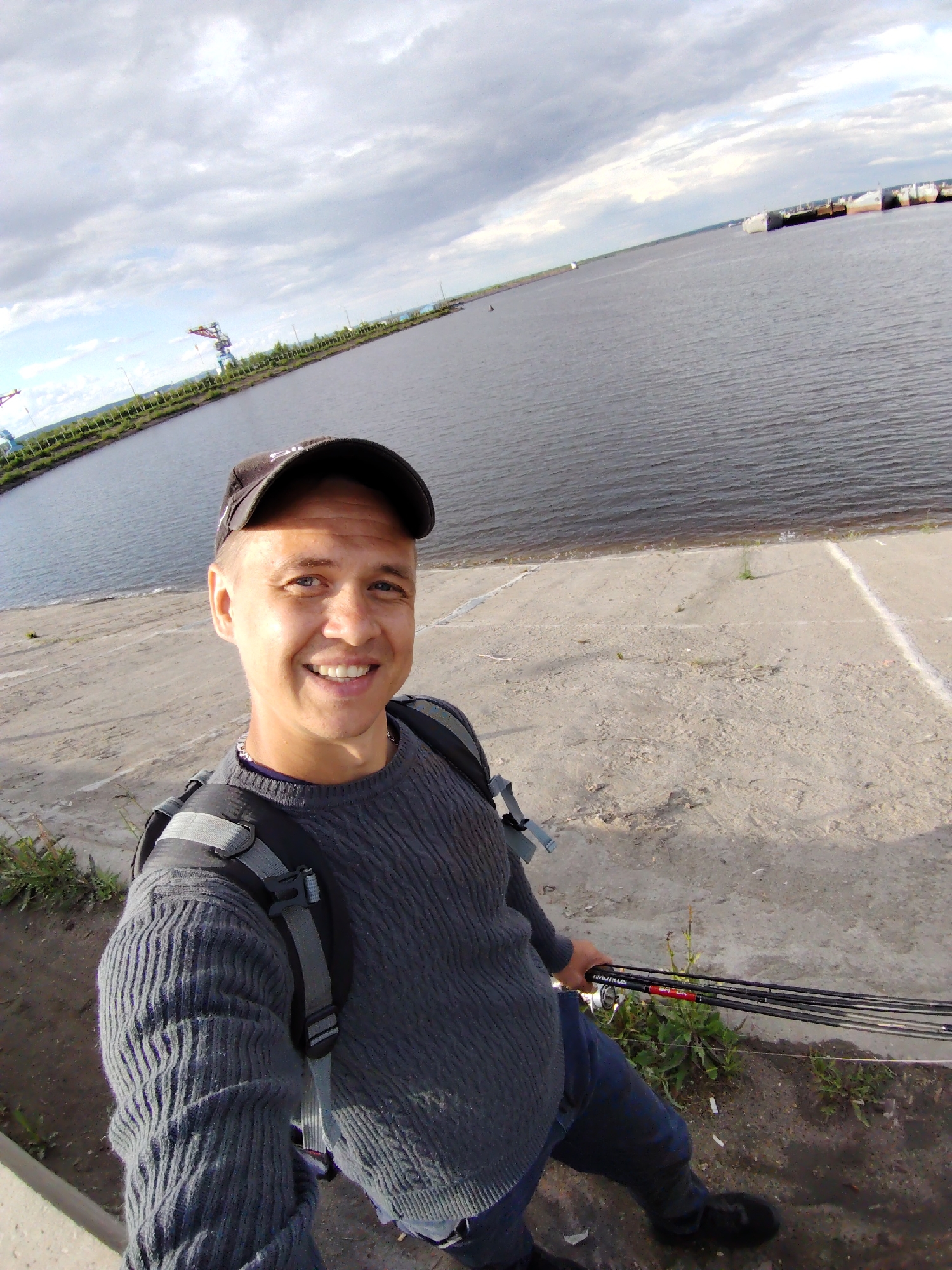 A man in a black baseball cap, gray sweater, and blue jeans stands on a concrete embankment, holding several fishing rods in his right hand. He is smiling and looking at the camera, presumably taking a selfie. He has a gray backpack on. Behind him is a large body of water, possibly a river or reservoir, with a shoreline visible in the distance. On the shoreline, there are structures that appear to be part of an industrial area, including cranes. The sky is overcast with gray clouds. The concrete embankment has some scattered vegetation. The location is likely the city of Chaikovsky, Russia, due to the presence of industrial structures and the body of water.