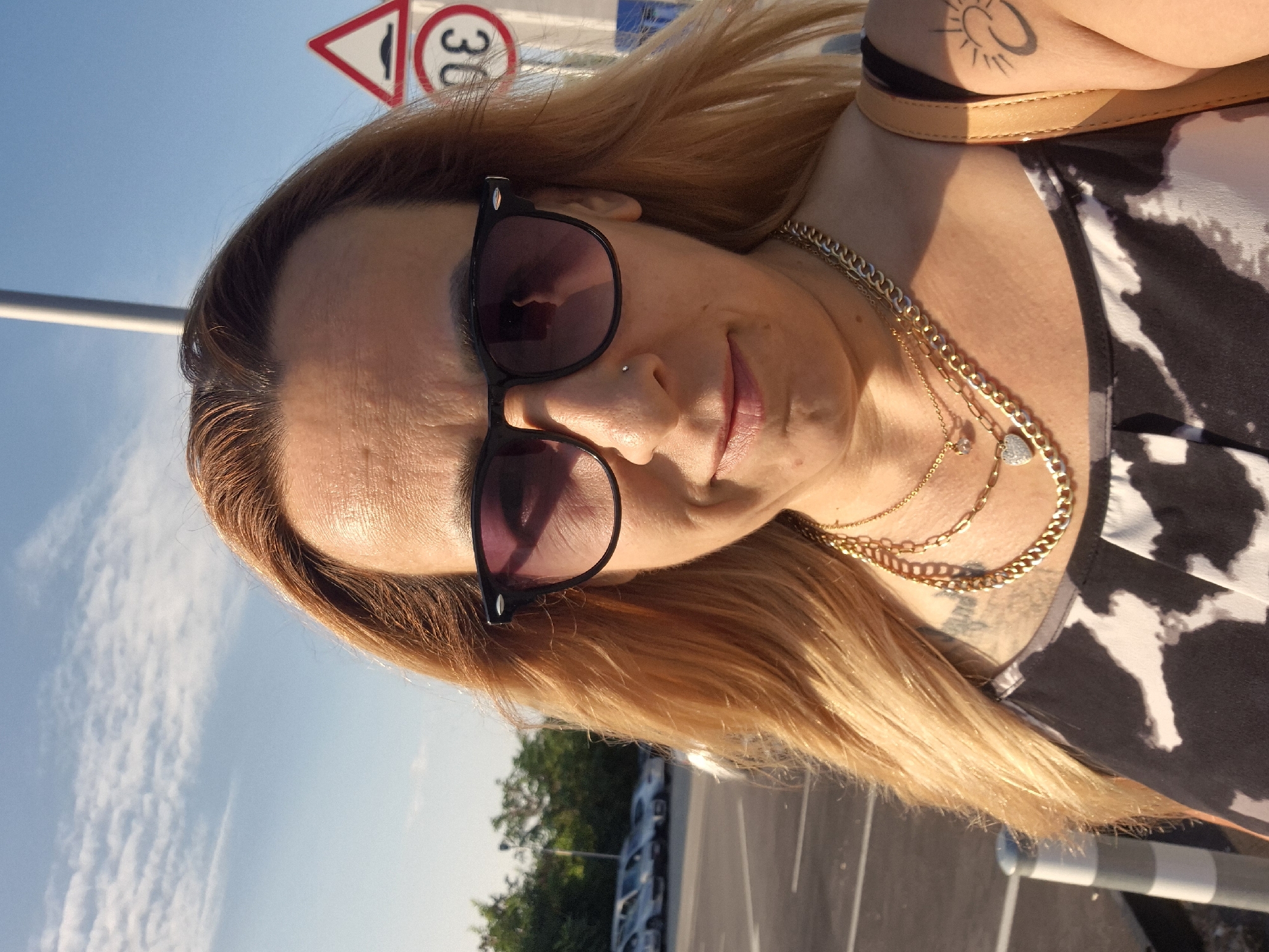 An outdoor, medium-shot, selfie-style photo captures a fair-skinned woman with long, blonde hair wearing black-rimmed sunglasses and a black-and-white patterned dress. She is adorned with multiple gold necklaces, one featuring a heart-shaped pendant and another with a small clear stone. She has a nose piercing and a tattoo on her left shoulder blade of a sun with rays.
Behind her, a paved parking lot is visible, with a row of cars parked in the distance. The background also shows trees and a bright blue sky with scattered clouds. Two traffic signs are present on the right side of the frame: a triangular sign with a speed bump symbol and a circular sign with a red border and the number '30' inside. The lighting suggests it is daytime, possibly late afternoon due to the warm tones.