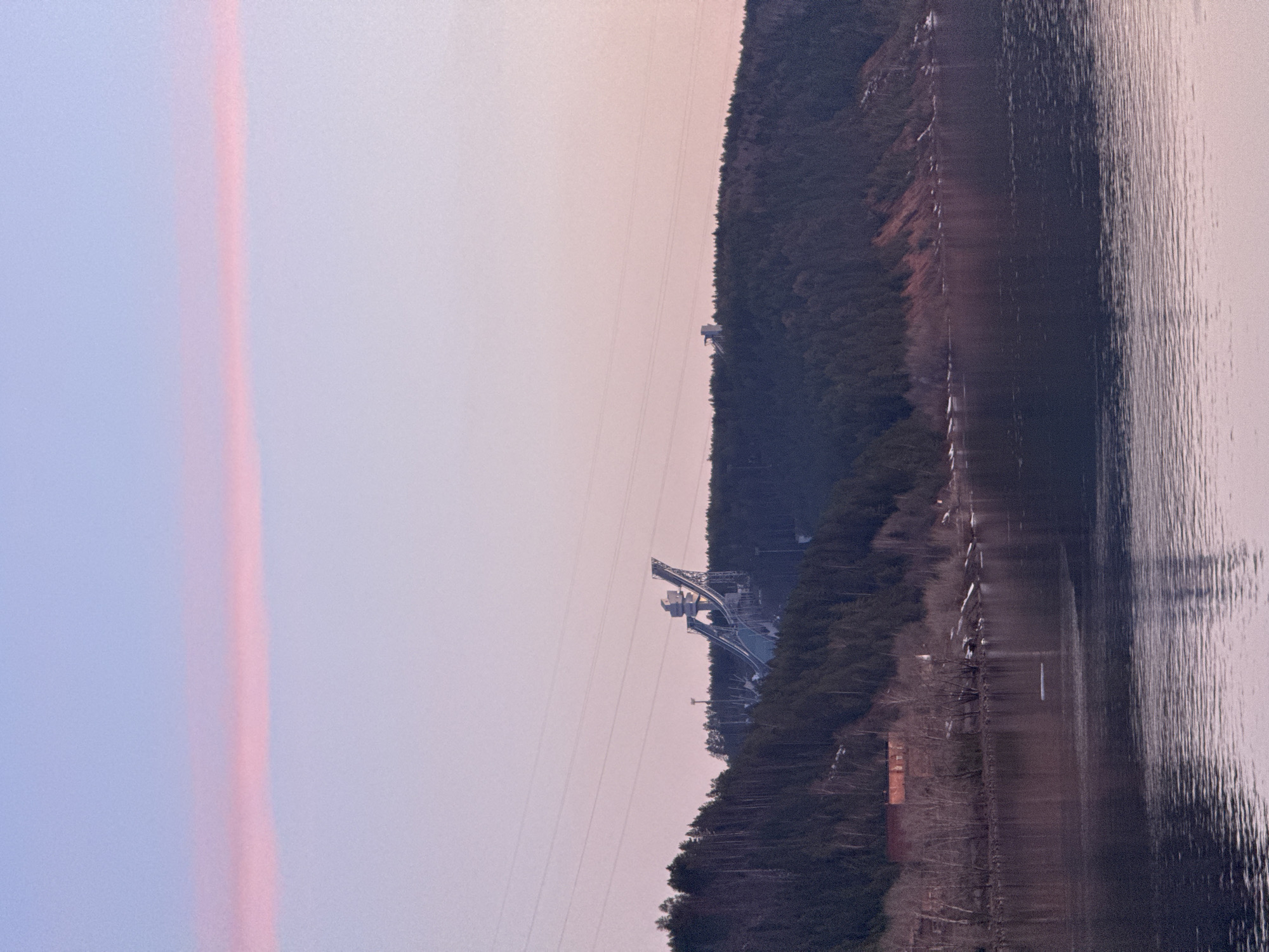 This panoramic image depicts a tranquil landscape in Chaikovsky, Russia, likely captured during dawn or dusk. The sky, which forms the upper portion of the scene, displays soft, diffused pastel colors ranging from light blue to purple and pink. A distinct, elongated reddish-pink atmospheric streak or cloud stretches horizontally across the sky on the left.
Below the sky, a dense, dark green forested bank runs horizontally through the middle ground. Dominating this forested area is a large, modern ski jump complex, with its prominent steel structures and multiple ramps, characteristic of the "Snezhinka" facility. Thin power lines are visible stretching across the sky, passing over the forest and near the ski jump.
The lower half of the image features a wide body of water, likely a river or lake, reflecting the soft hues of the sky and showing subtle ripples on its surface. The scene combines natural beauty with significant human-made sporting infrastructure, under a calm, ethereal sky. No specific activities or visible text are discernible in the image.