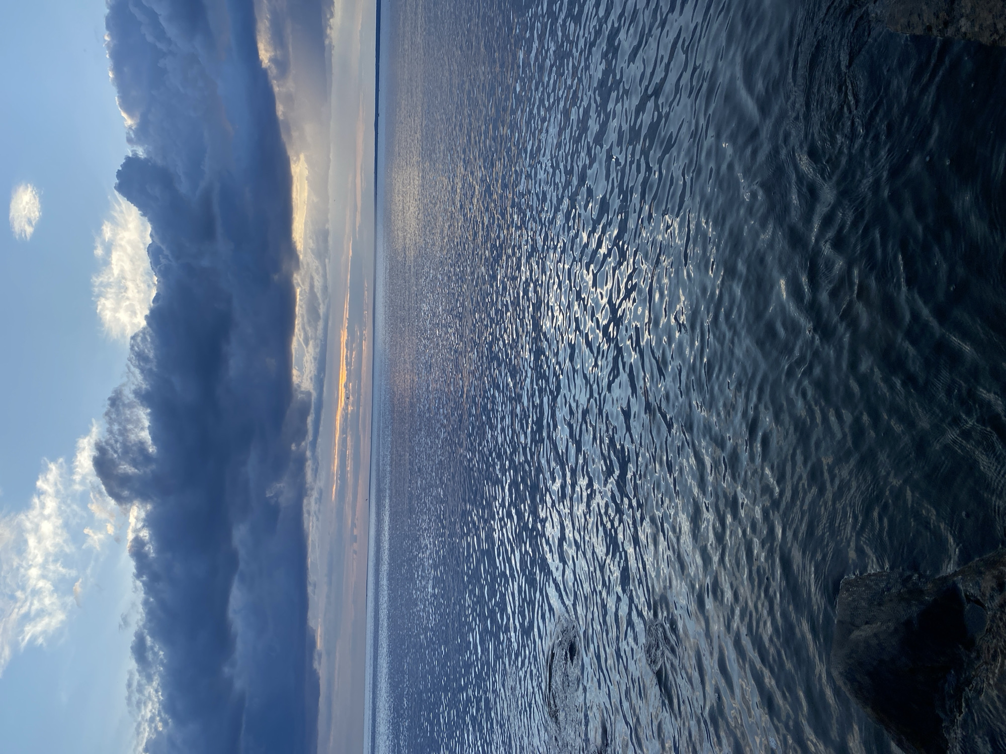 A wide-angle vertical shot captures a serene body of water under a dramatic, cloud-filled sky, likely taken at dusk or dawn. The foreground features dark, rough-textured rocks partially submerged on the left side, with the rippling water extending to the horizon. The water surface reflects the muted light, creating a shimmering effect. Dark, heavy clouds dominate the upper portion of the image, with lighter, wispy clouds interspersed. A sliver of warm light from the setting or rising sun peeks through a gap in the clouds near the horizon. The scene evokes a sense of tranquility and the vastness of nature. This vista is consistent with the coastal areas of Sestroretsk, Russia, known for its Gulf of Finland shoreline.