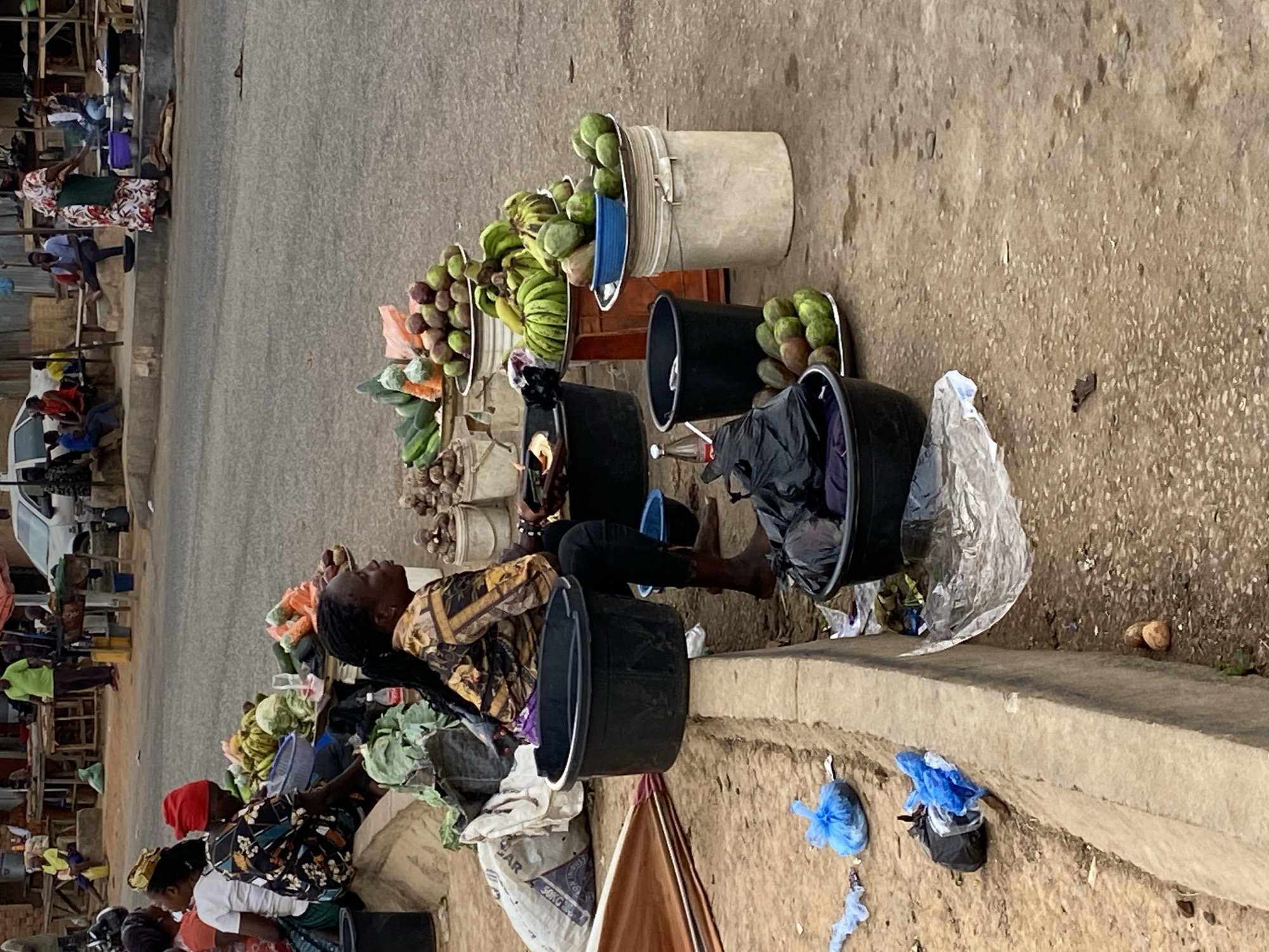 This image captures an outdoor market scene in Bukuru, Nigeria, showing several individuals engaged in vending activities. The primary focus is on the right side (when rotated to the correct vertical orientation), where vendors display fresh produce along a dirt path adjacent to a paved road.
Several people are visible. In the foreground, a woman with braided hair, wearing a patterned gold, black, and brown top, sits on a concrete curb, looking to her right, away from the camera. Her legs are tucked to the side. Nearby, another woman wearing a red head covering and a patterned dress sits further down the curb, facing left. In the background, more individuals are seen sitting or standing near stalls and a white vehicle.
The ground-level market area is filled with various fruits and vegetables arranged in plastic buckets and bowls. These include green mangoes, bananas, and leafy greens resembling cabbage or lettuce. Black and white plastic buckets serve as containers for the goods. Scattered around are plastic bags, some blue, and a partial view of a brown umbrella or awning lies on the ground. A plastic bottle is visible near one of the black buckets. A large white sack, partially visible, displays the text "...AR 50KG," suggesting it contains a significant quantity of goods.
The scene depicts a typical bustling daytime market, with bright, natural light indicating a clear day. Vendors are waiting with their produce, creating a lively commercial atmosphere characteristic of a street market.