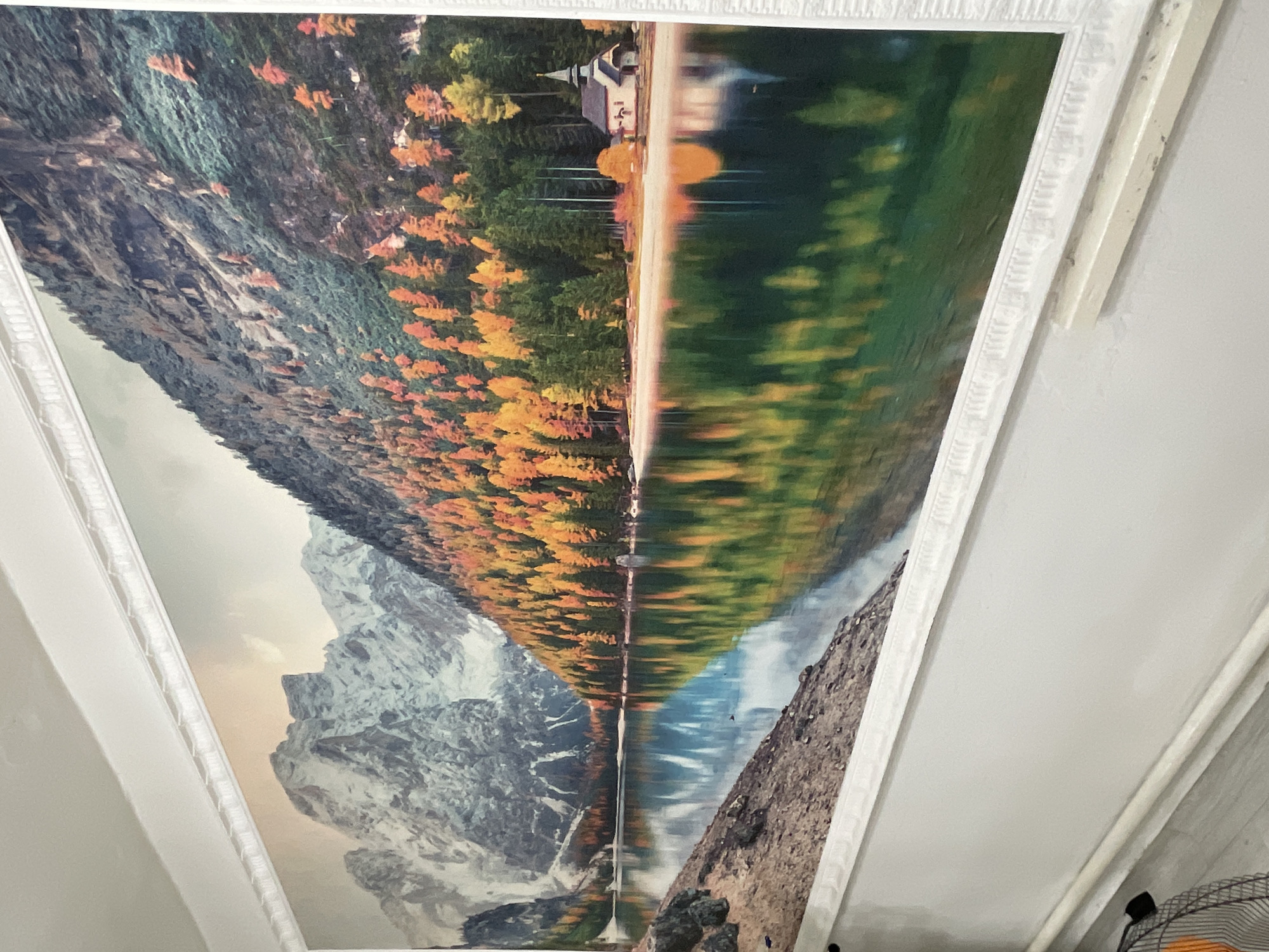 A panoramic mural depicting a scenic mountain lake landscape in autumn is mounted on a white wall. The mural features snow-capped mountains in the background, a forest with trees in vibrant fall colors of orange, yellow, and green, and a calm lake reflecting the scenery. On the right side of the lake, a small white building with a dark roof and a steeple, resembling a church or chapel, is visible. The mural is framed by decorative white molding. Below the mural, the wall transitions to a lighter shade, and a light-colored baseboard runs along the bottom. A white pipe is affixed to the wall above the baseboard. In the lower left corner of the image, a portion of a black and orange fan is visible. The overall scene suggests an indoor setting, likely a room in a residential or commercial space, with the mural serving as a decorative element. There is no indication of the specific location in Bishkek City, Kyrgyzstan.