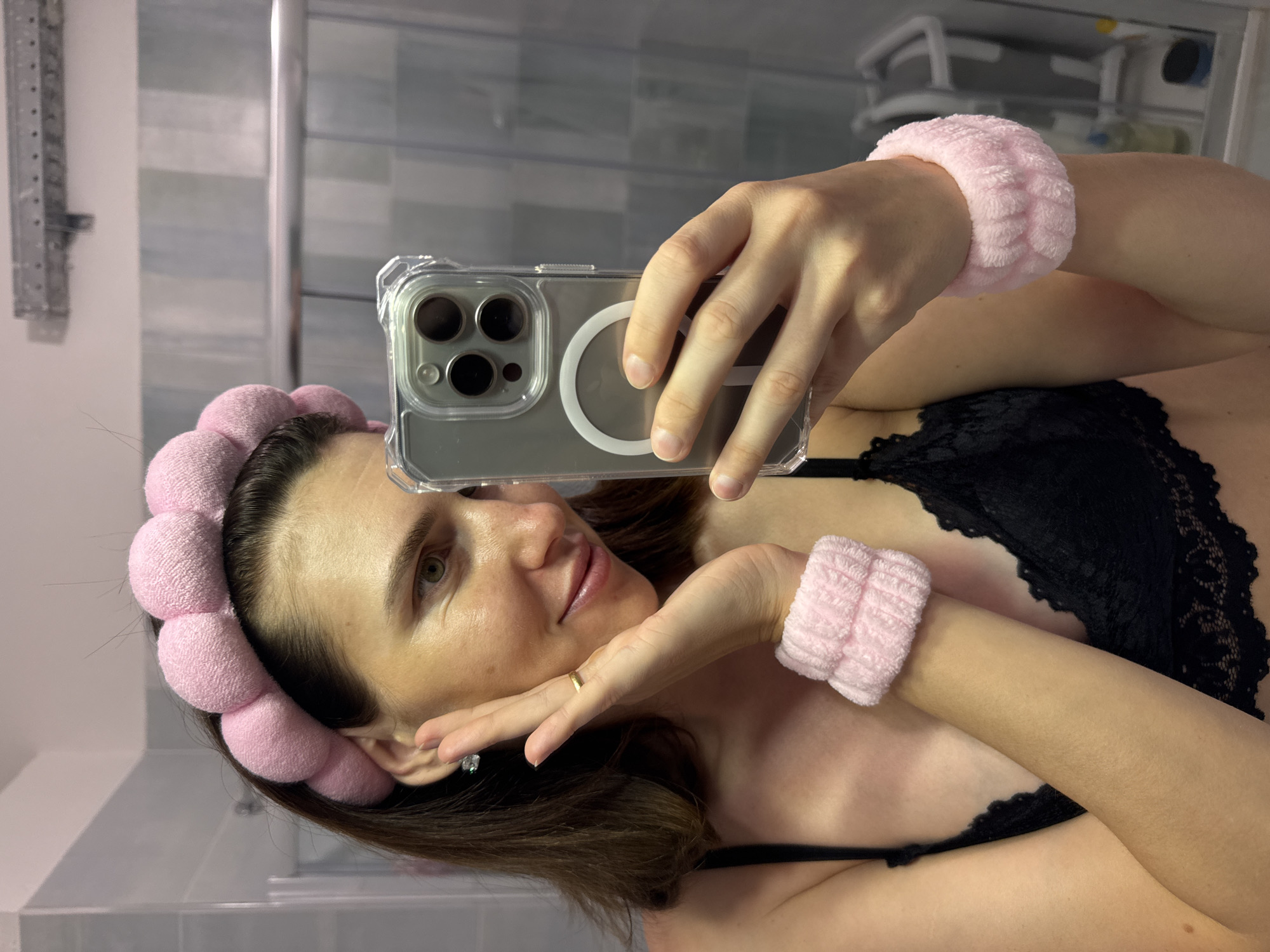 A woman with dark brown hair, wearing a black lace bra, a pink fluffy headband, and pink fluffy wristbands, holds a smartphone to take a selfie in a bathroom. Her right hand is on her cheek, and she is smiling. The bathroom features light-colored tiled walls and a shower enclosure. The image does not contain any identifiable landmarks or features that would indicate its location in the city of Isola delle Femmine, Italy.