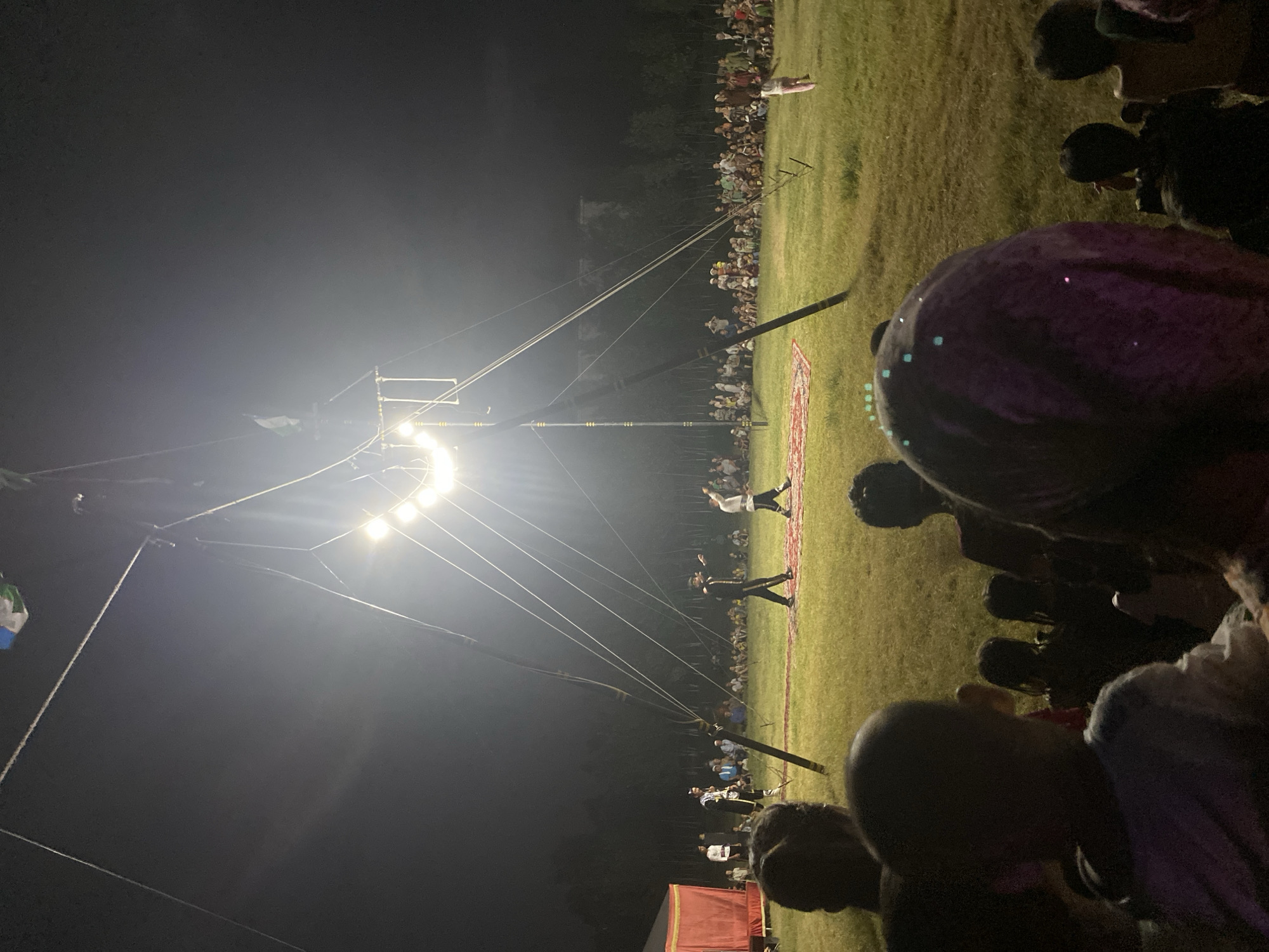 An outdoor circus performance is underway at night in Arbagish, Uzbekistan, illuminated by bright stage lights strung from a tall, tripod-like structure. The performance area is a grassy field with a red carpet laid out for the performers. Three individuals are visible on the carpet, dressed in performance attire. One, in the center, appears to be leading the performance, gesturing with his arms. To his left and right, two other performers are also engaged in the act, one of whom is walking with a raised hand. A large audience, comprising adults and children, is seated on the grass, watching the show. In the background, a tent with a red curtain is set up on the left side of the field. The overall scene depicts a community gathering for an evening of entertainment in a rural or park-like setting within Arbagish.