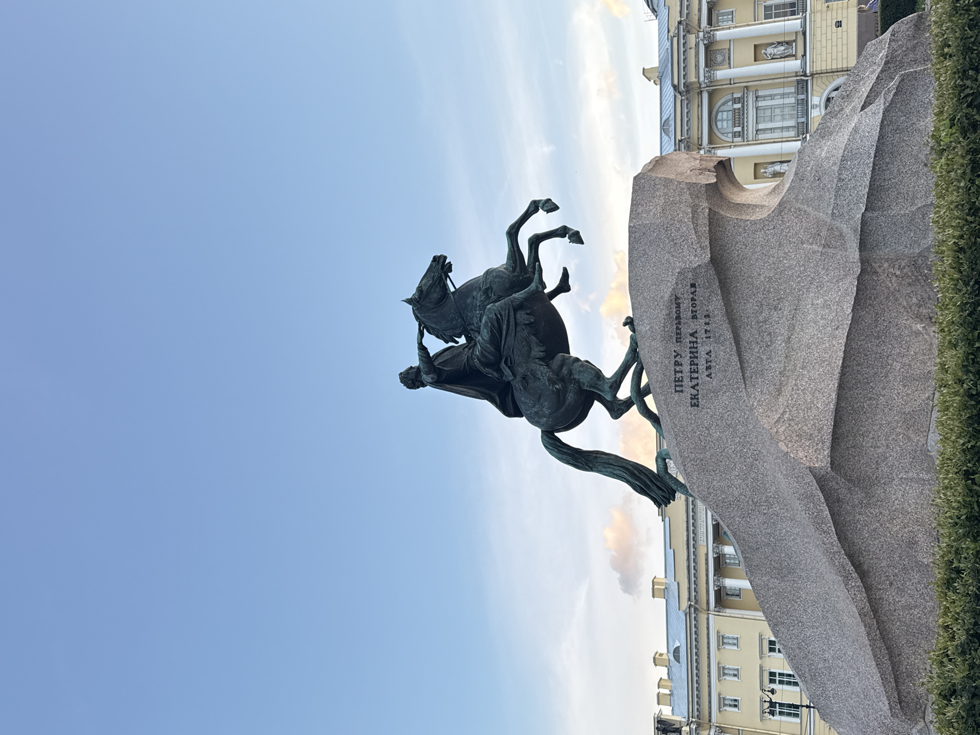 The image shows the Bronze Horseman, a famous equestrian statue of Peter the Great, located in Senate Square in Saint Petersburg, Russia. The statue is made of bronze and depicts Peter the Great on horseback, with his right hand extended forward. The horse is rearing up on its hind legs, appearing to be climbing a large rock. The statue is mounted on a massive granite pedestal known as the Thunder Stone. In the background, there is a large, yellow neoclassical building with columns and arched windows, likely part of the surrounding historic architecture of Senate Square. The sky above is clear and blue with some light clouds. The scene is captured from a low angle, emphasizing the grandeur of the statue and its setting.