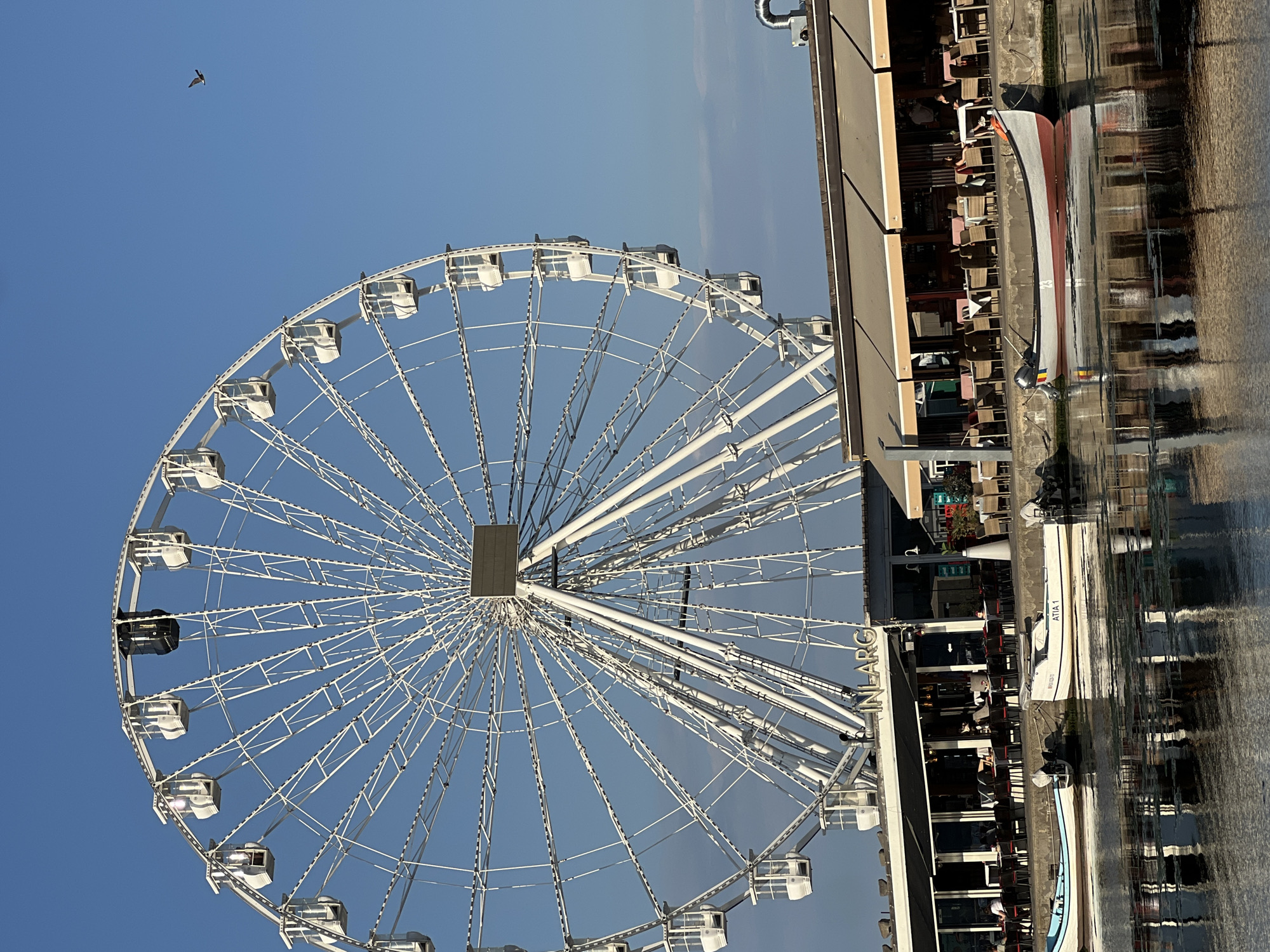 A large white Ferris wheel dominates the frame against a clear blue sky. The Ferris wheel has numerous passenger cabins, some of which appear to be white and one that is black. Below the Ferris wheel, a waterfront restaurant with outdoor seating is visible. Tables and chairs are arranged under beige awnings. Several small boats are moored in the water in front of the restaurant. The water reflects the scene above, including the Ferris wheel and the restaurant. The image appears to be taken from a slightly low angle, looking up towards the Ferris wheel and across the water. The text "INLARG" is visible on the facade of the building. The location is Mamaia, Constanța, Romania.