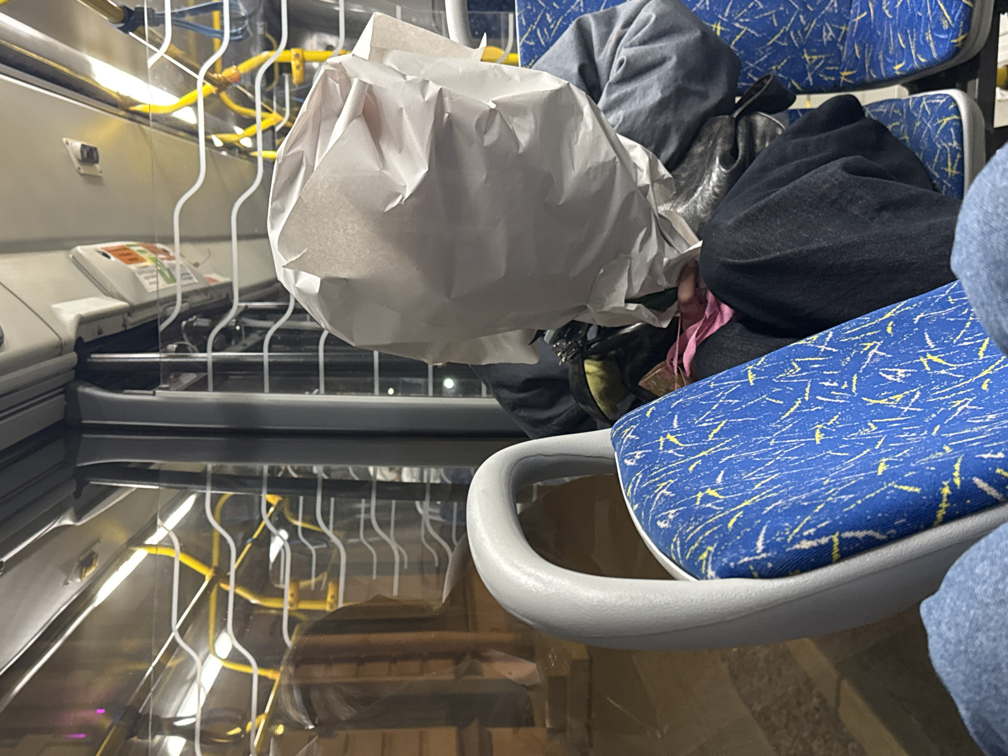 The image captures a scene inside public transportation, likely a bus or tram, in Saint Petersburg, Russia, based on the presence of typical interior elements. An occupied blue seat with yellow and white accents and a gray handle is in the foreground. A crumpled white paper bag dominates the central part of the image, alongside gray and black clothing items placed on the adjacent seat, indicating passenger belongings. Reflected in the glossy floor is the interior of the vehicle, including white vertical poles, yellow railings, and a control panel or fare validator. The floor's reflective quality suggests a recently cleaned or wet surface.