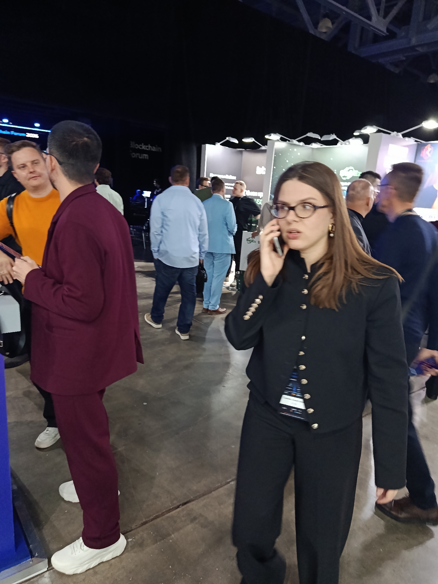 The image shows a bustling scene, possibly at a conference or trade show, in a large indoor venue. A woman with glasses and dark hair is talking on her cell phone. She is wearing a black jacket with gold buttons and black trousers. Her attire suggests a professional or business-oriented event.
Several other people are visible in the background. A man in a maroon suit is walking in the opposite direction. There are several men wearing light colored shirts and another man is wearing an orange shirt.
Signs, possibly indicating the name of the event, are partially visible in the background. The lighting is artificial. The floor appears to be a light colored concrete.
There is no definitive evidence in the image to suggest the location is in Khimki, Russia. The location cannot be verified using this image.