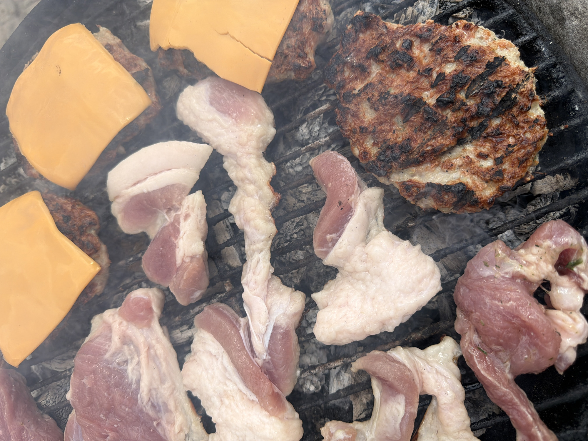The image shows a grill with various food items cooking on it. There are several pieces of raw pork with visible fat marbling. Additionally, there are cooked burger patties, some with slices of orange cheese melting on top. The grill itself is made of metal bars, and there is smoke rising from the cooking food. The background is blurred, but it appears to be an outdoor setting, though it is not possible to confirm the location as Lesichovo, Bulgaria, from the image alone.
