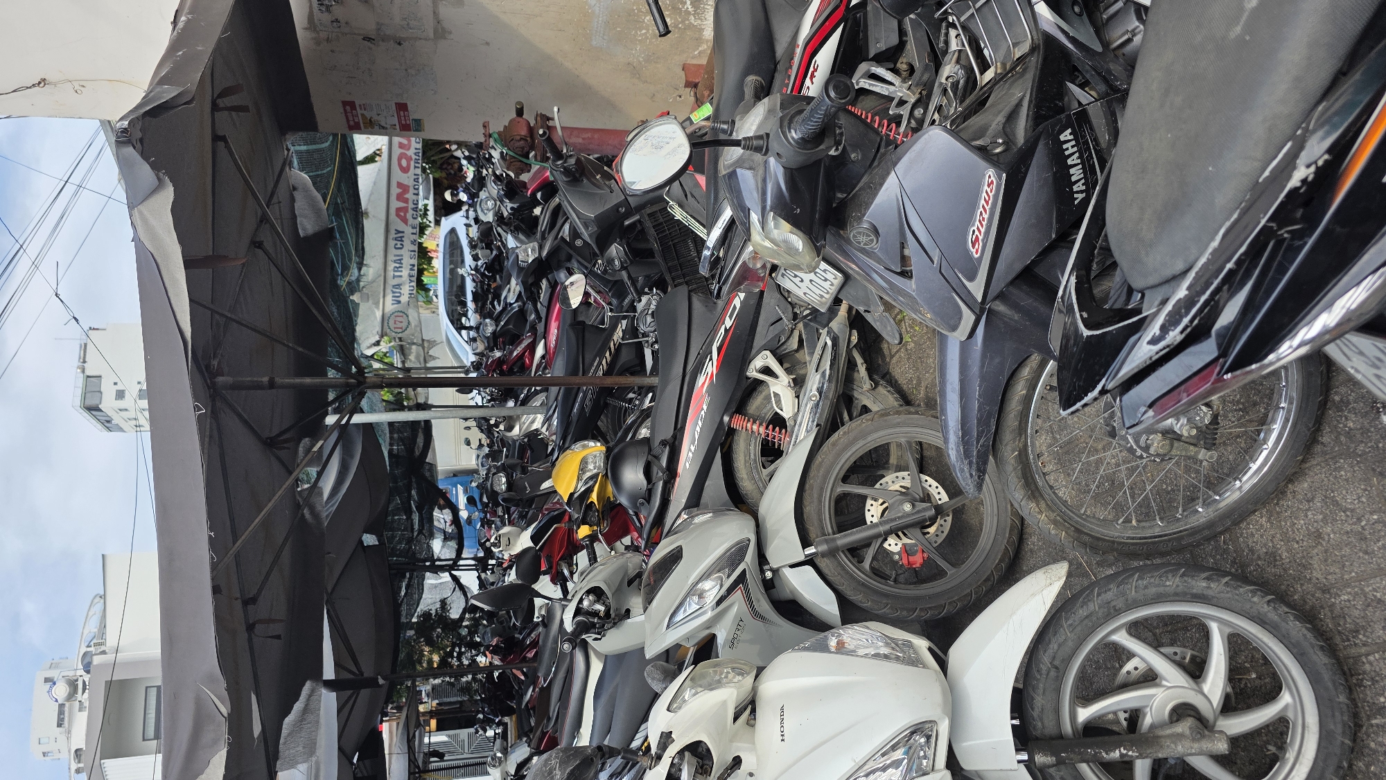 This image captures a dense outdoor parking area for motorcycles and scooters in Nha Trang Ward, Vietnam, under bright daylight. Numerous two-wheeled vehicles are packed closely together, indicating a busy urban environment, possibly near a market or commercial establishment.
The scene is predominantly filled with a variety of motorcycles and scooters. Visible brands and models include "HONDA," "YAMAHA," "SIRIUS," "SPORTY," and "BLADE SPO." Many of the vehicles show signs of regular use, with some visible scuffs and dirt. Partial license plate numbers, such as "140.95" and "79," are also discernible on some of the parked bikes.
Overhead, several large, dark grey market umbrellas are open, providing shade for the parked vehicles and potentially an adjacent vendor stall. In the background, beyond the umbrellas and motorbikes, parts of buildings are visible against a bright, clear sky.
A large white sign with Vietnamese text is partially visible through the gaps in the umbrellas and vehicles. The text on this sign reads: "VỰA TRÁI CÂY AN QUY" (Fruit Stall An Quy), followed by "CHUYÊN SỈ & LẺ CÁC LOẠI TRÁI C" (Specializing in Wholesale & Retail of Fruits). This suggests a fruit stall or market operates nearby. No specific human activity is clearly depicted, but the sheer volume of parked vehicles implies high foot traffic and commerce in the area.