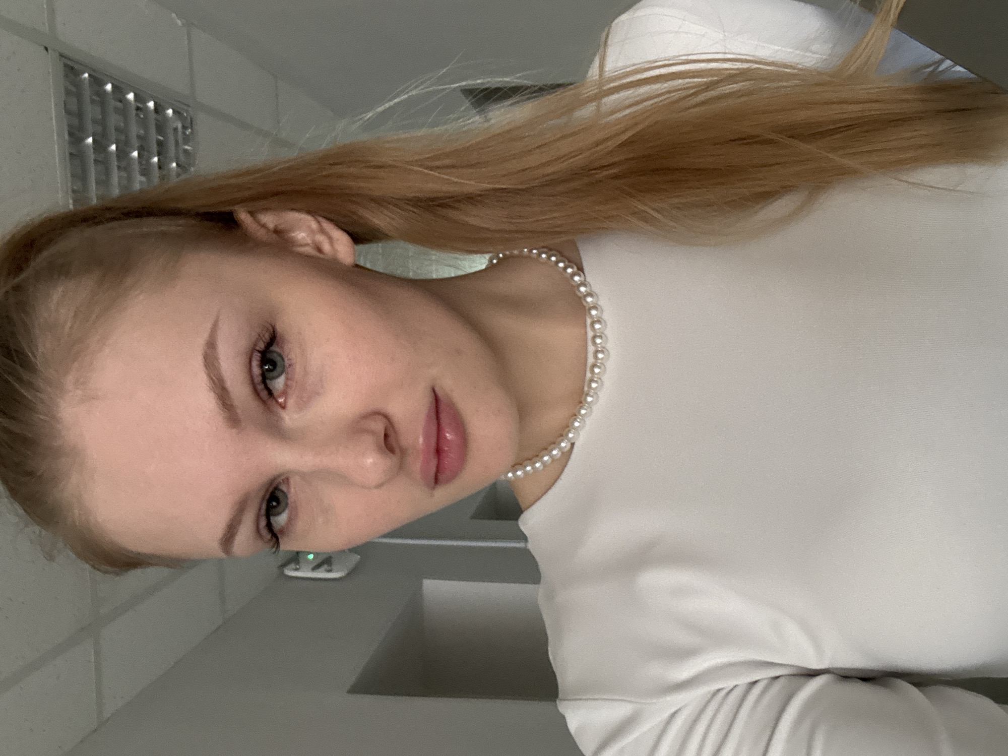 The media file presents an indoor portrait of a young woman. She has fair skin, light-colored eyes (appearing blue or green), and long, straight blonde or light brown hair styled in a high ponytail. She is wearing a white long-sleeved top and a pearl necklace. Her head is slightly tilted, and she gazes directly at the camera with a neutral and calm expression.
The background features plain light gray walls and a ceiling. A rectangular light fixture or air vent is visible on the ceiling in the upper left portion of the image. Further back, a small white electronic device with a green indicator light is affixed to the wall. A light gray surface, possibly a desk or countertop, is visible in the lower right foreground. The lighting appears to be artificial and cool-toned, indicative of an indoor environment such as an office or public space. No text is visible within the image. The location context provided for this image is Perm, Russia.
