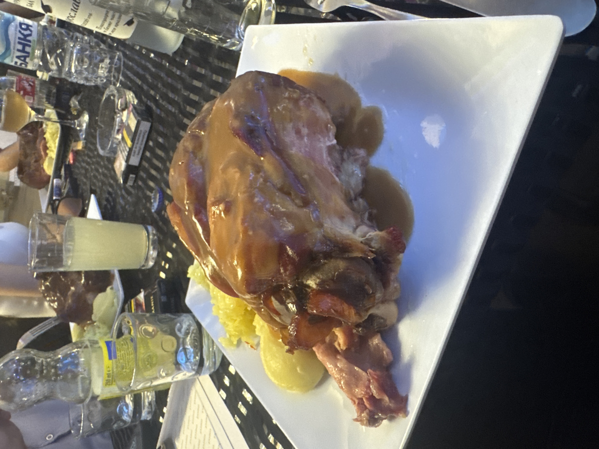 The image depicts a prepared food dish on a white square plate. The main element is a large piece of roasted meat, likely pork, covered in a glossy sauce. Around the meat, there are sides that could be potatoes and sauerkraut. The meal is set on a table with a dark, patterned surface, likely in a restaurant setting. Surrounding the plate are various bottles and glasses, some filled with liquid, and what appears to be a cigarette packet. The location is Pazardzhik, Bulgaria.