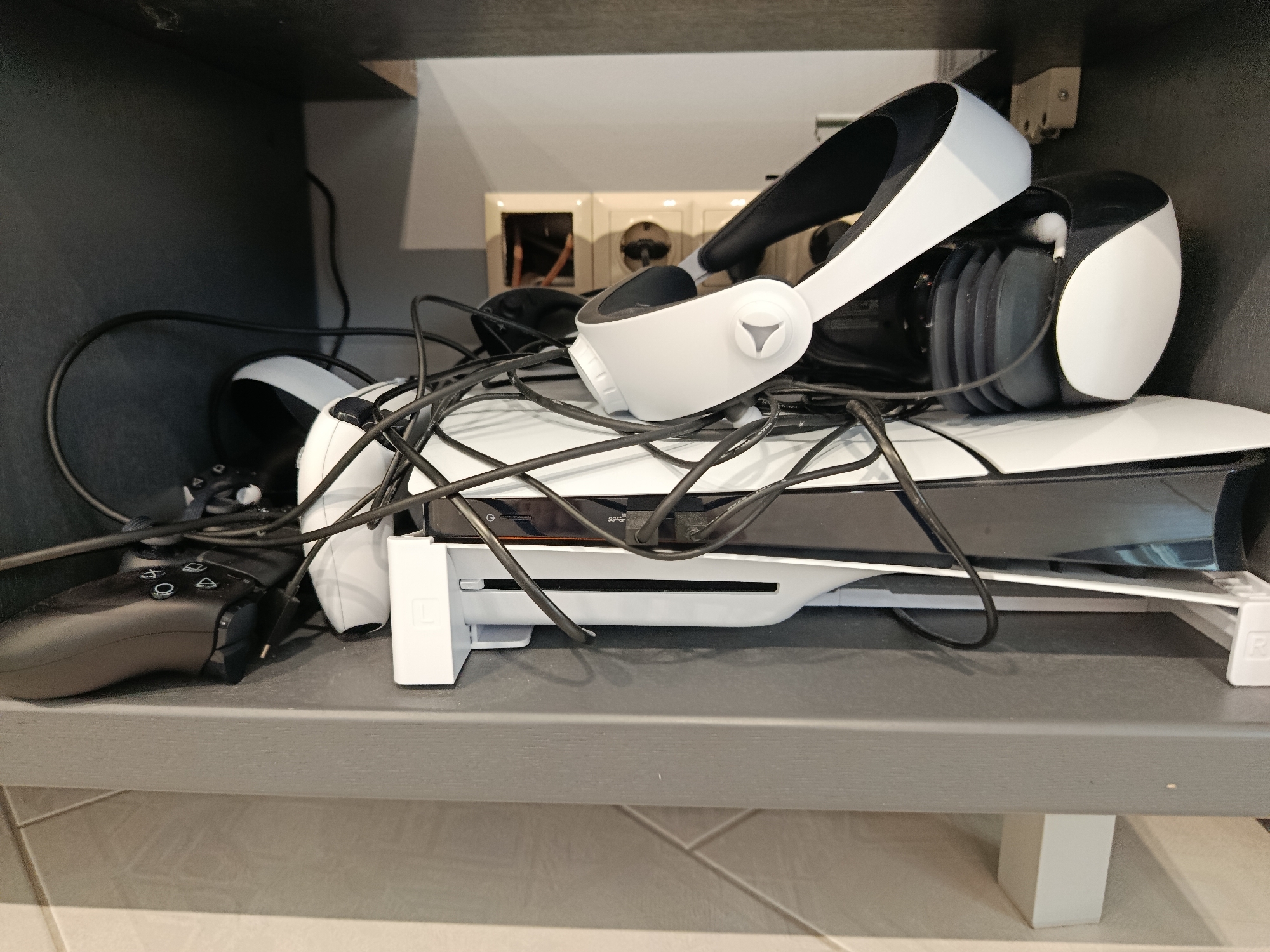 A PlayStation 5 console, a VR headset, and a controller are neatly organized on a shelf. The VR headset rests on top of the console, with the controller placed to the left. The scene appears to be inside a home, with a gray shelf and a dark background. The lighting is dim, suggesting an indoor environment. There are no people visible in the image, and no specific location in Taganrog, Russia can be identified.