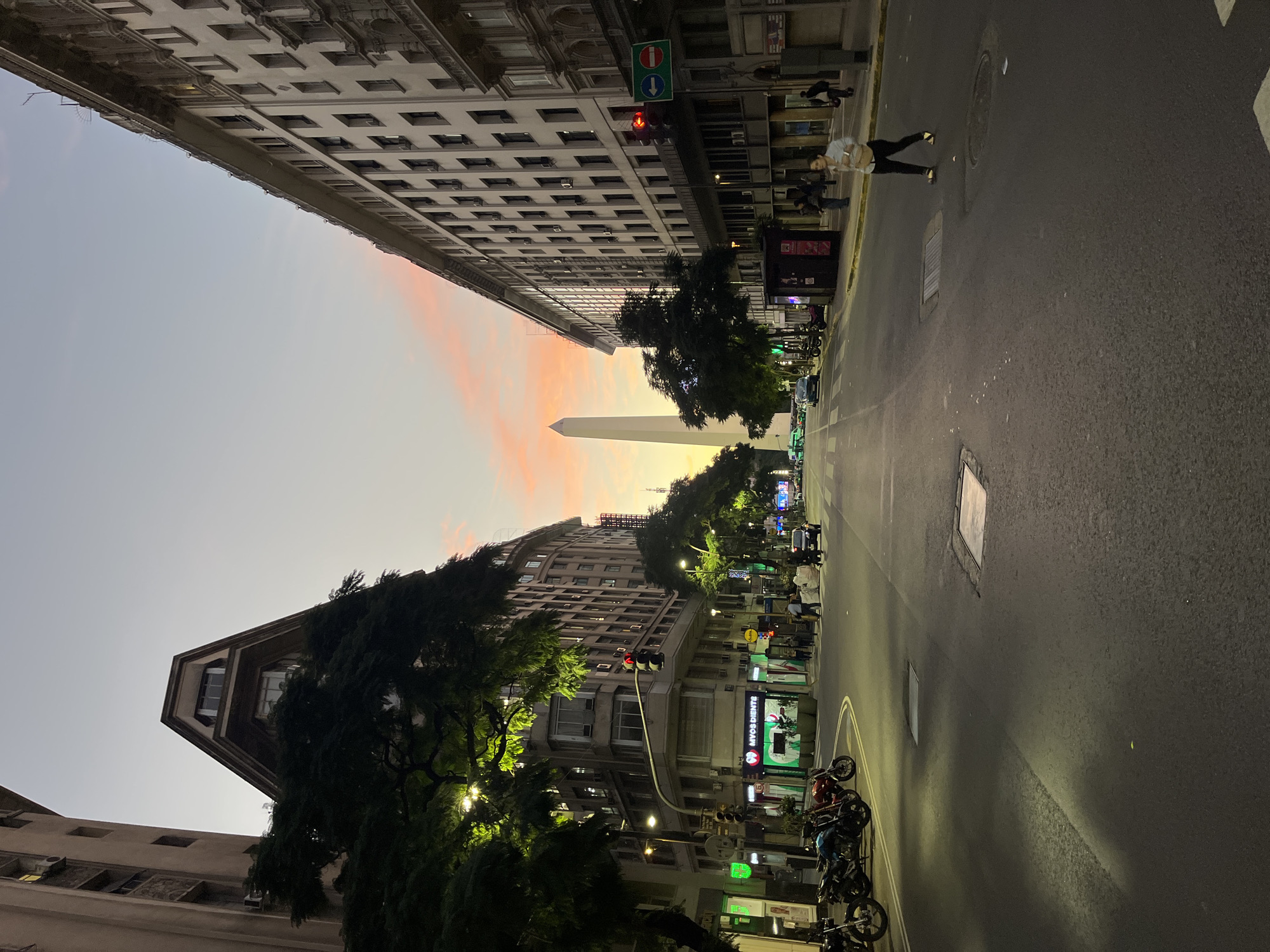 This media file presents a captivating urban scene in Buenos Aires, Argentina, captured during the twilight hours. The sky displays a beautiful gradient, transitioning from a soft, light blue at the top to warm orange and pink tones near the horizon, strongly suggesting either a sunset or sunrise. The natural light fades, while artificial illumination from streetlights, building windows, and shop signs begins to glow, casting a gentle glow on the surroundings.
The image looks down a wide city street, which appears to have multiple lanes, flanked on both sides by tall, multi-story buildings featuring numerous windows. These structures exhibit a classic urban architectural style. In the distance, perfectly framed by the converging buildings, stands the prominent white Obelisco de Buenos Aires, an iconic monument, silhouetted against the colorful sky.
On the street level, a few pedestrians are visible on the sidewalks, and one person wearing a light top and dark pants is seen walking or standing on the right side of the road. Several motorcycles are parked along the right curb. Lush green trees are interspersed along the street, adding natural elements to the urban landscape, with some foliage illuminated by nearby lights. A red traffic light is visible on the right, accompanied by a green sign with white arrows indicating "up" and "down" traffic directions. On a building on the right, a shop sign clearly displays "MYOS DIENTE". The scene exudes a tranquil and atmospheric mood as the city transitions from day to night.