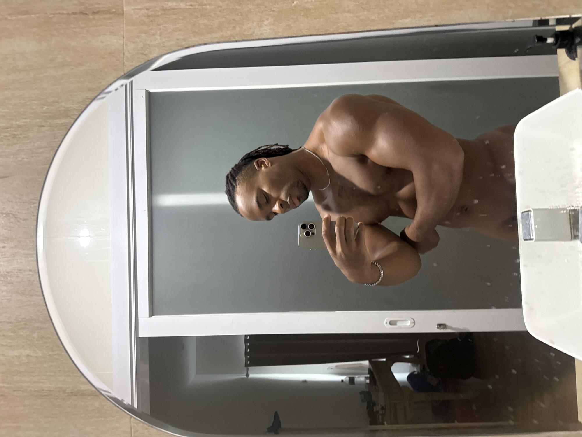 The image shows a shirtless man taking a selfie in a bathroom mirror. The mirror is oval-shaped with a white frame, and reflects the man's torso and head, as well as a glimpse of the bathroom interior including a white sink and cabinet beneath the mirror. The man has dark skin, braided hair, and is wearing a silver necklace and bracelet. He holds a smartphone in his left hand. The bathroom wall appears to be tiled in a light brown color. The location is Unknown, Unknown.