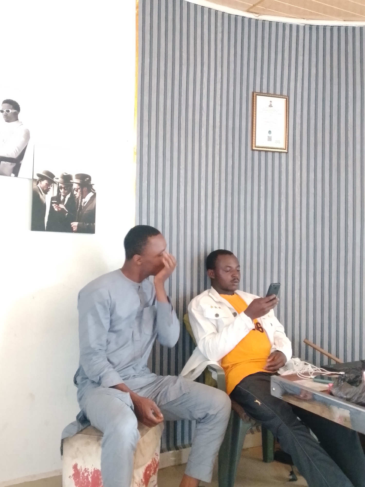 Two adult males are seated indoors in a casual setting, likely a waiting area or informal business space. The room features a plain white wall on the left and a vertically striped wall with grey and white bands on the right. The ceiling above the striped wall is curved and appears to be made of light-colored wood panels.
On the left, a man wearing a light blue or grey traditional long-sleeved garment (possibly a kaftan or similar attire) sits on a low, repurposed drum or stool, which has some red and white markings on its base. He is turned slightly to his right, looking away from the camera, with his right hand covering part of his face, possibly indicating thought, weariness, or an action like a yawn or cough.
On the right, another man is seated on a green plastic chair, looking down intently at a black mobile phone in his hands. He is wearing an orange t-shirt, a white long-sleeved jacket adorned with three small gold stars on the left shoulder, and dark trousers. A table is positioned to his right, with white cables and some dark items visible on its surface.
The white wall section on the left displays two framed black-and-white pictures. The upper one shows a man in sunglasses and a light-colored top. The lower one depicts three men in hats and suits, one appearing to be looking at a phone. A single framed certificate or document hangs on the striped wall, though its text is not legible. The overall ambiance is relaxed, with both individuals engaged in separate activities.