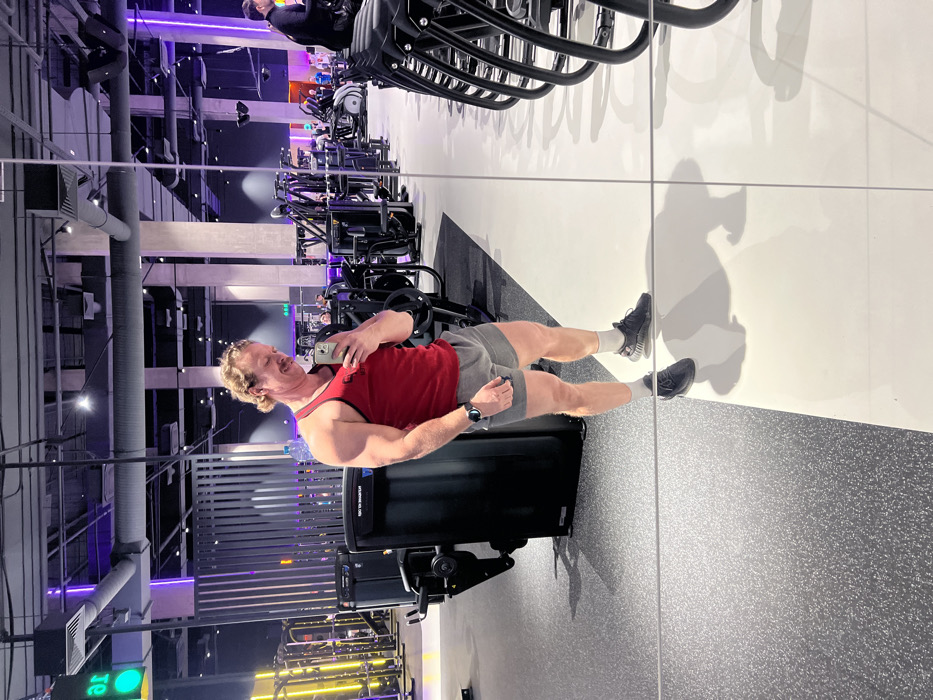 The image captures a selfie taken in what appears to be a modern gym or fitness center, reflected in a large mirror. The primary subject is a man with a light complexion and light brown or reddish-blonde hair styled in a bun or ponytail at the back of his head, along with a mustache. He is wearing a red sleeveless tank top, grey athletic shorts, white socks, and dark athletic shoes. A smartwatch is visible on his left wrist. He is holding a light-colored smartphone (possibly silver or white) in his right hand, pointing it towards the mirror to take the photo, and appears to be smiling or making a lighthearted expression. His left arm is flexed, showcasing his bicep.
The man stands on a grey speckled rubber floor, which transitions into a clean white wall in the background of his reflection. A thin vertical line in the mirror indicates a seam or edge. His reflection shows a strong silhouette on the white wall behind him. Behind the man, on the gym floor, is a black piece of exercise equipment, possibly a hip thrust machine or similar strength training apparatus, with a dark blue or purple accent on its side.
The gym itself is spacious, with high ceilings and industrial-style exposed beams and ventilation ducts. The lighting is a mix of ambient white light and distinctive purple and yellow LED lighting accents, particularly on the walls and around equipment. In the background, numerous pieces of gym equipment are visible, including exercise bikes, weight machines, and possibly free weights, arranged in rows. Another person can be seen in the upper left corner of the image (in the reflection), seated on an exercise machine. The overall atmosphere suggests a well-lit and contemporary fitness facility.
No clear text is fully legible, but a green illuminated sign with inverted text (likely due to the reflection) is partially visible in the far background on the left side of the gym.