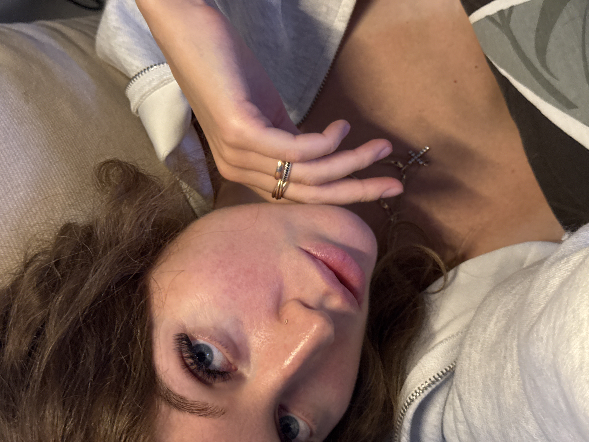 An overhead close-up shot captures a person, likely female, lying down indoors in Chaikovsky, Russia, looking directly at the camera. Her face occupies the lower half of the frame, tilted slightly. She has light-colored skin, prominent eyelashes, and blue eyes. A small stud piercing is visible on her left nostril, and her lips are slightly parted, revealing a natural pink hue. Her brown, wavy hair frames her face and spreads out on a light-colored, textured fabric surface, possibly a pillow or couch.
Her right hand is raised towards her face and neck, with several rings worn on her fingers; one appears to be a golden wraparound design, and another is a band with dark embellishments. A silver-colored necklace with a small cross pendant, possibly adorned with stones, rests on her upper chest/neck area. She is wearing a light grey or white hooded jacket or sweatshirt with a visible zipper detail on the front. In the background, to the right, a darker patterned fabric, possibly part of another cushion or bedspread, is visible. The scene suggests a relaxed, casual, and intimate moment, with the person directly engaging the viewer. There is no visible text.