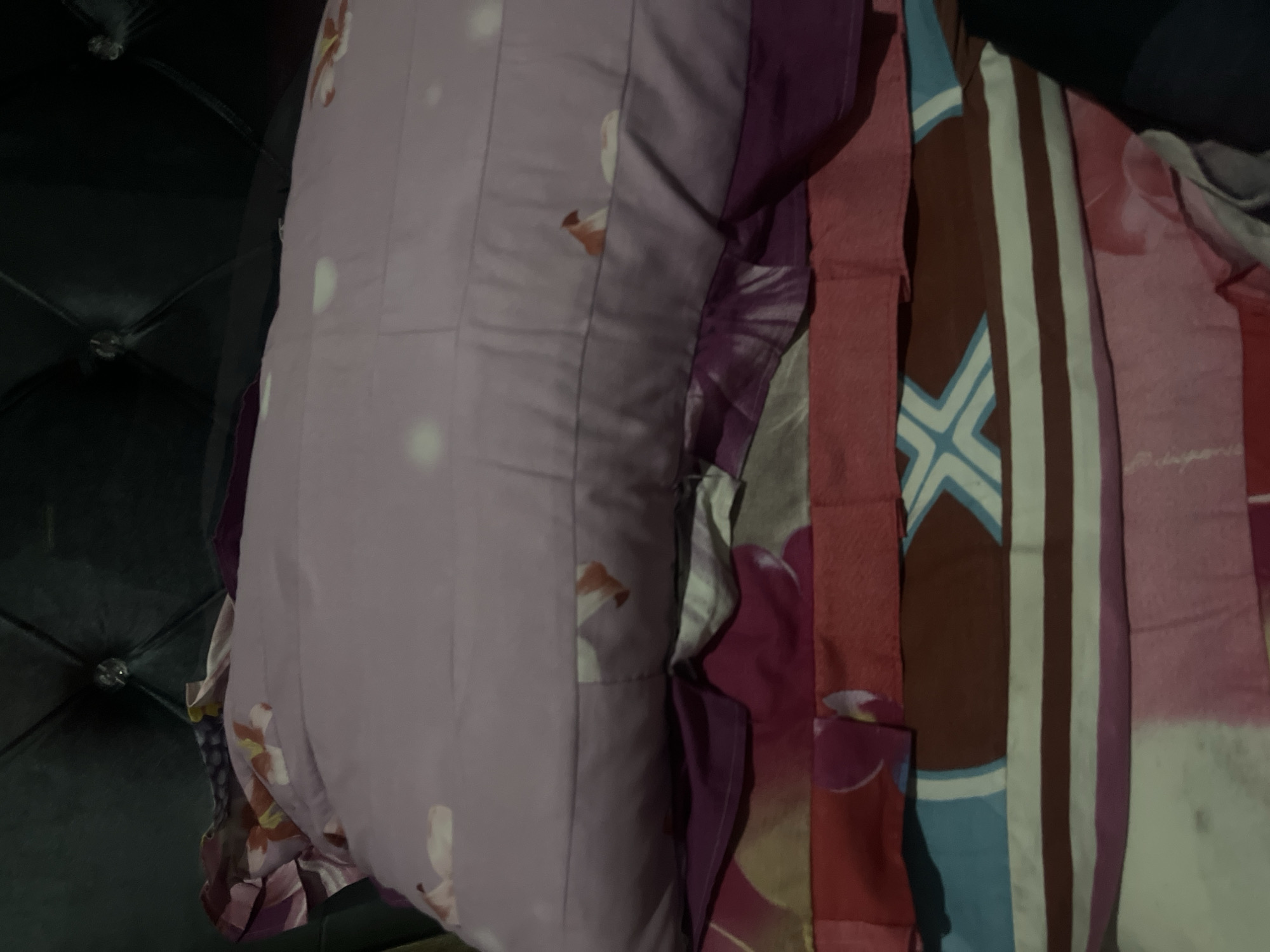A stack of folded bedding, including blankets and pillows, is arranged on a bed. The bedding exhibits a variety of colors and patterns. The uppermost item is a light purple pillow with faint white polka dots and floral accents in shades of pink and orange. Beneath this pillow, several layers of fabric are visible. A layer of dark purple fabric is next, followed by a layer with a bold floral pattern in shades of pink and yellow. Below this, there is a pink layer, and further down, a layer with a repeating geometric pattern featuring white, blue, and brown stripes. The very bottom layer appears to be white with a pink floral print. The bedding is positioned in front of a dark, tufted headboard, possibly made of leather or a similar material, with visible decorative buttons. The lighting in the image is dim, suggesting it was taken indoors. The scene is devoid of any individuals. This scene is located in Kebumen, Indonesia.
